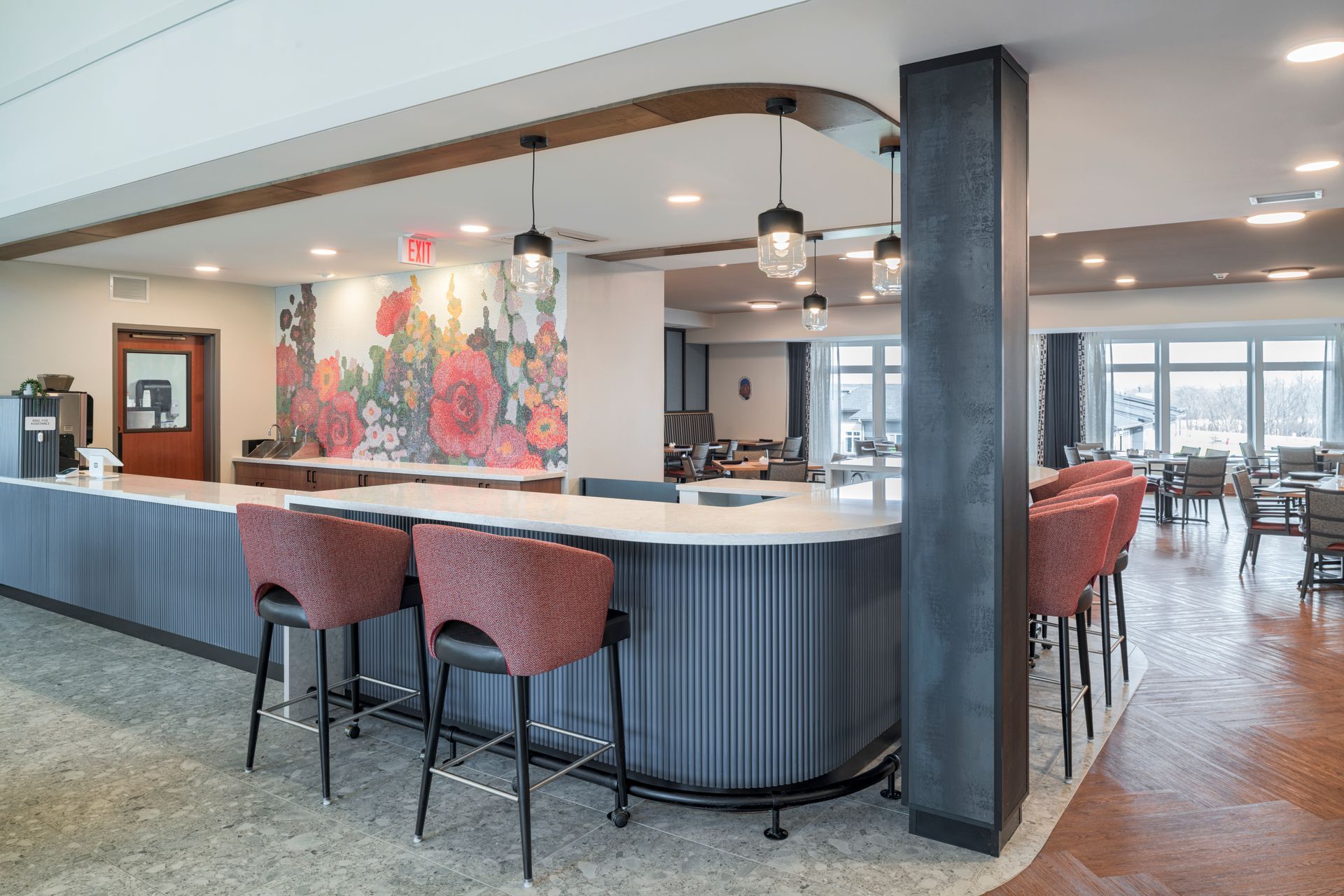 A contemporary bar area with a curved counter, red stools, floral wall mural, and warm lighting in a lobby setting.