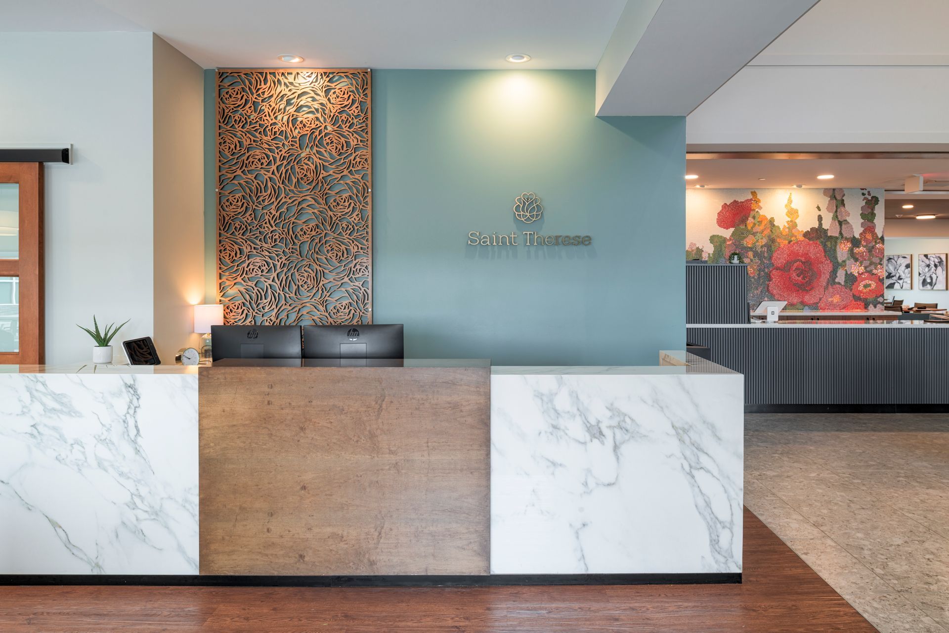 A reception desk with a white marble and wood facade, situated in a hotel lobby with a floral mural in the background.