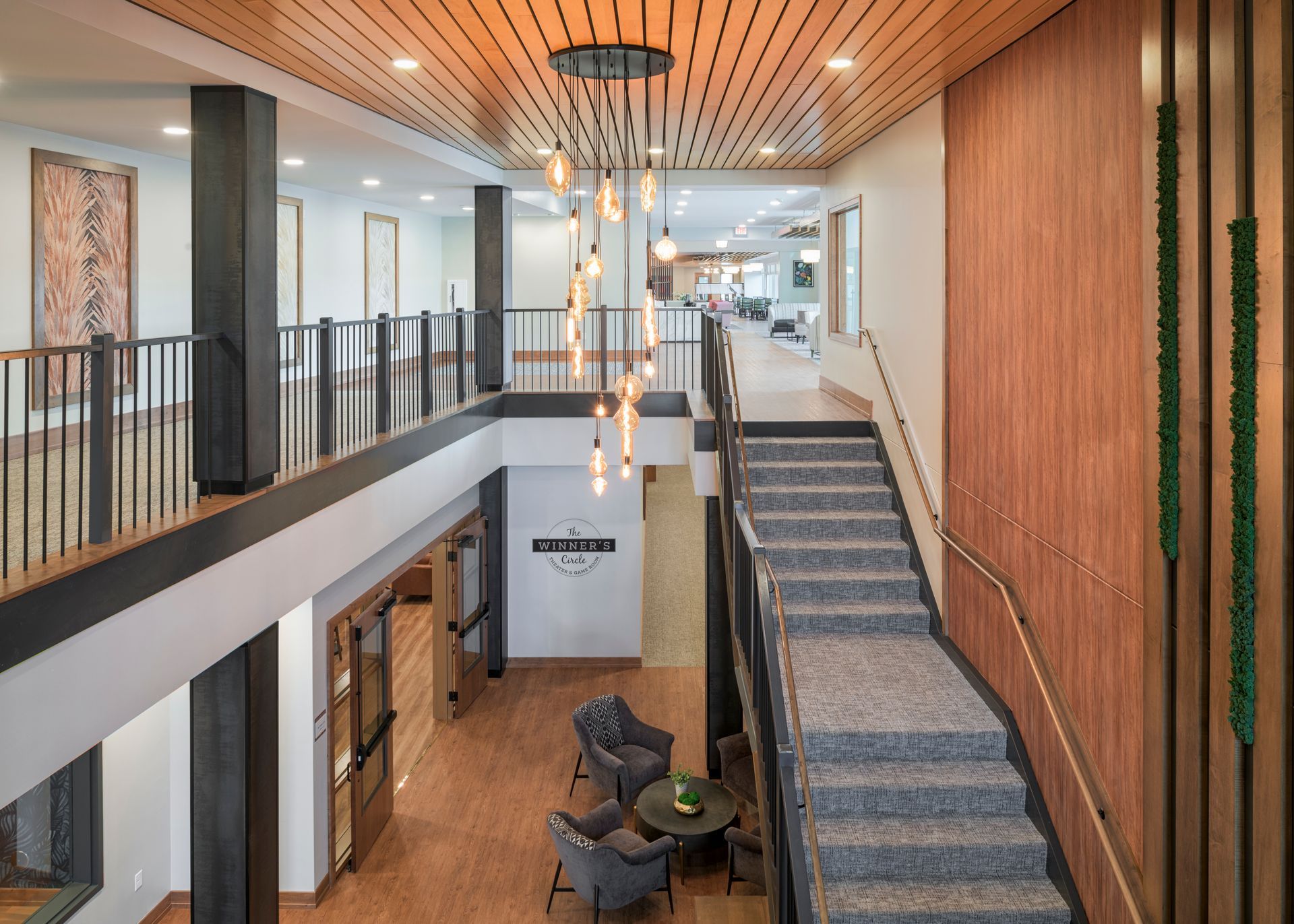 An indoor view of a multi-level lobby with a grand staircase, wooden ceiling, pendant lighting, and a small lounge area.