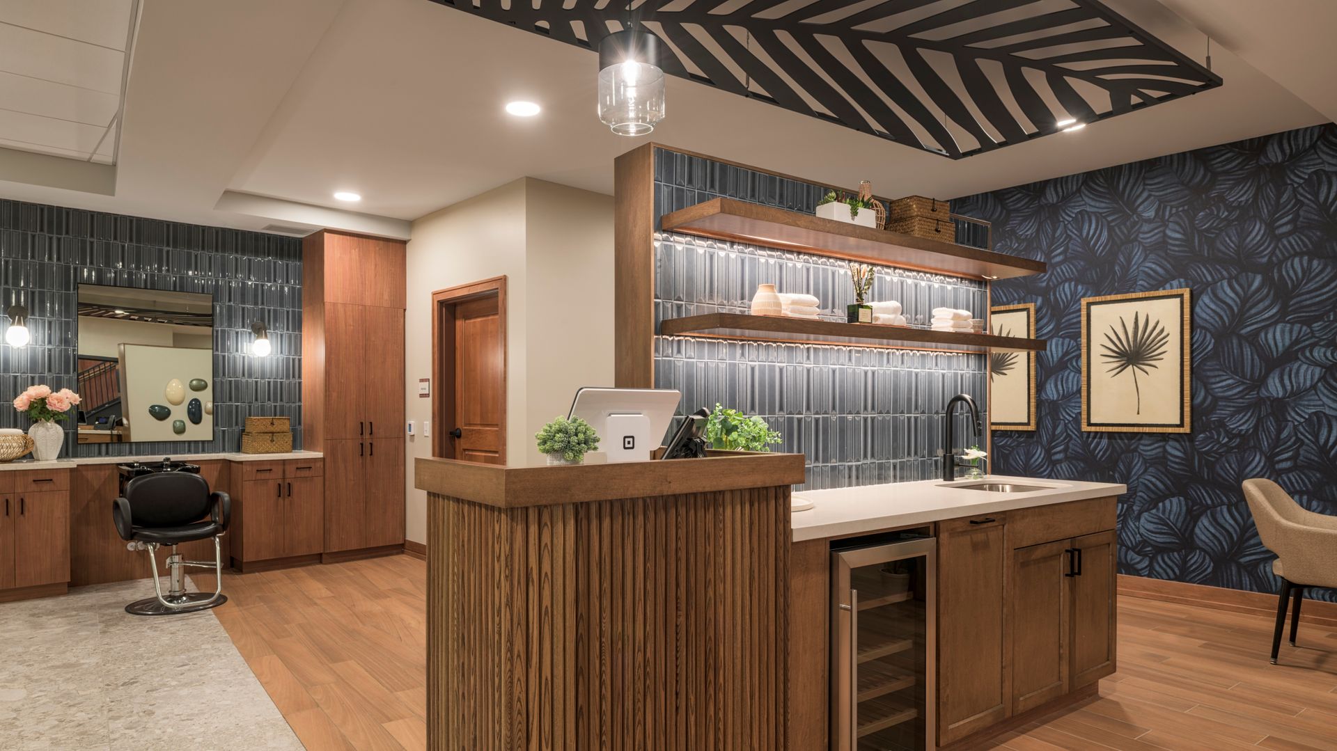 A modern salon or cafe interior featuring a wooden service bar, shelves with glassware, floral wallpaper, and a salon chair.