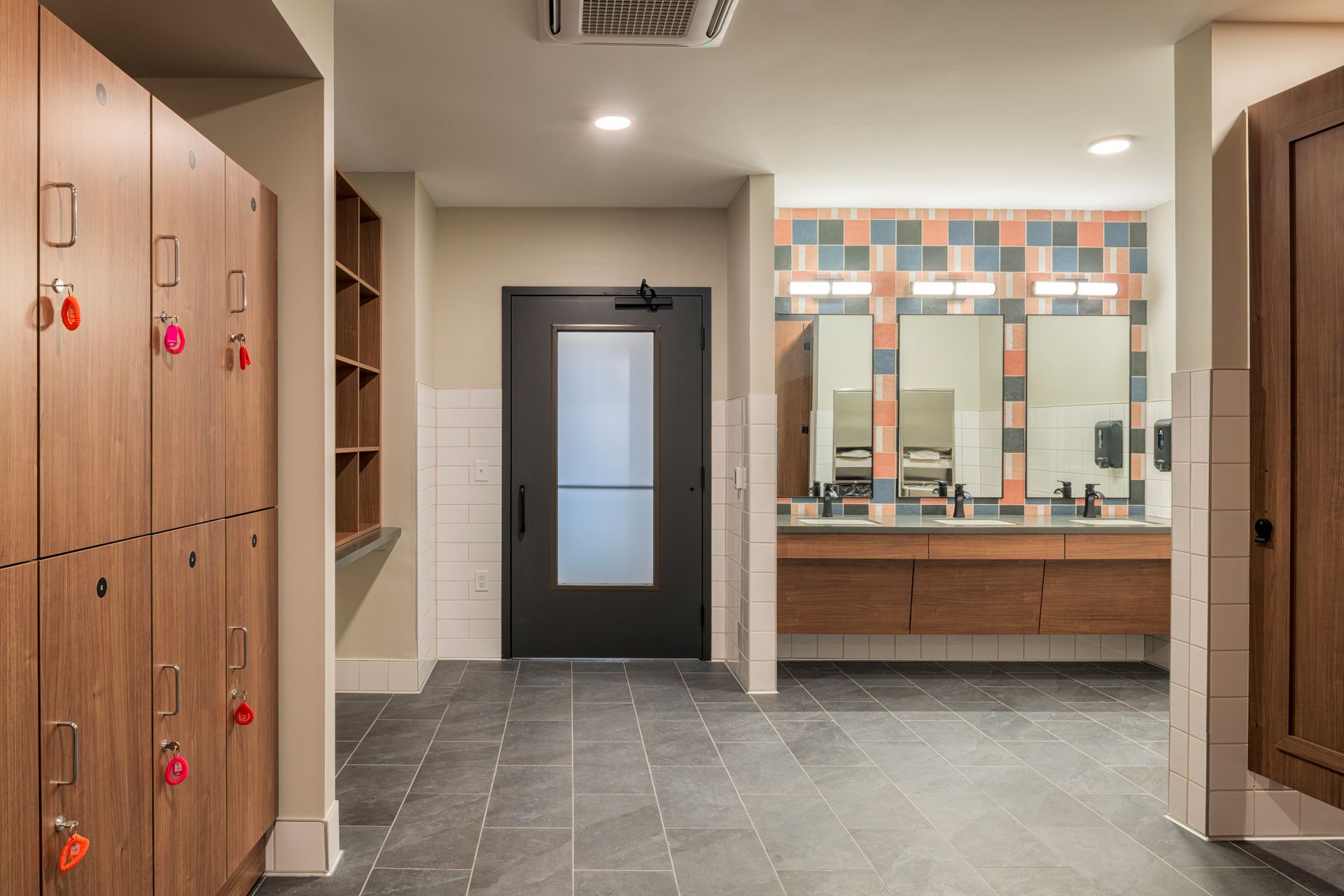 Locker room with wooden lockers on the left, a dark door in the center, and a vanity with mirrors and tiled walls.