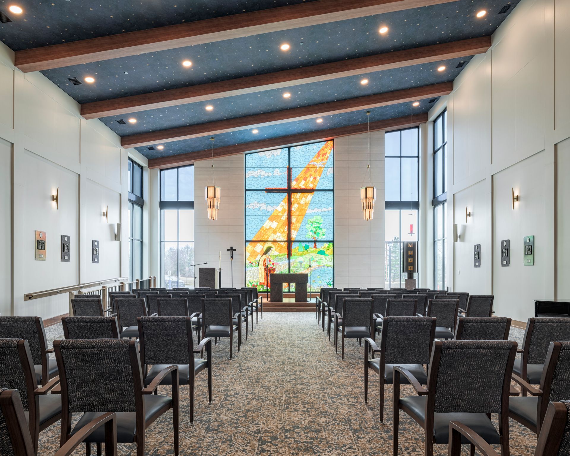 Interior of a chapel featuring rows of chairs facing a large stained-glass window depicting a cross under a star-lit ceiling.