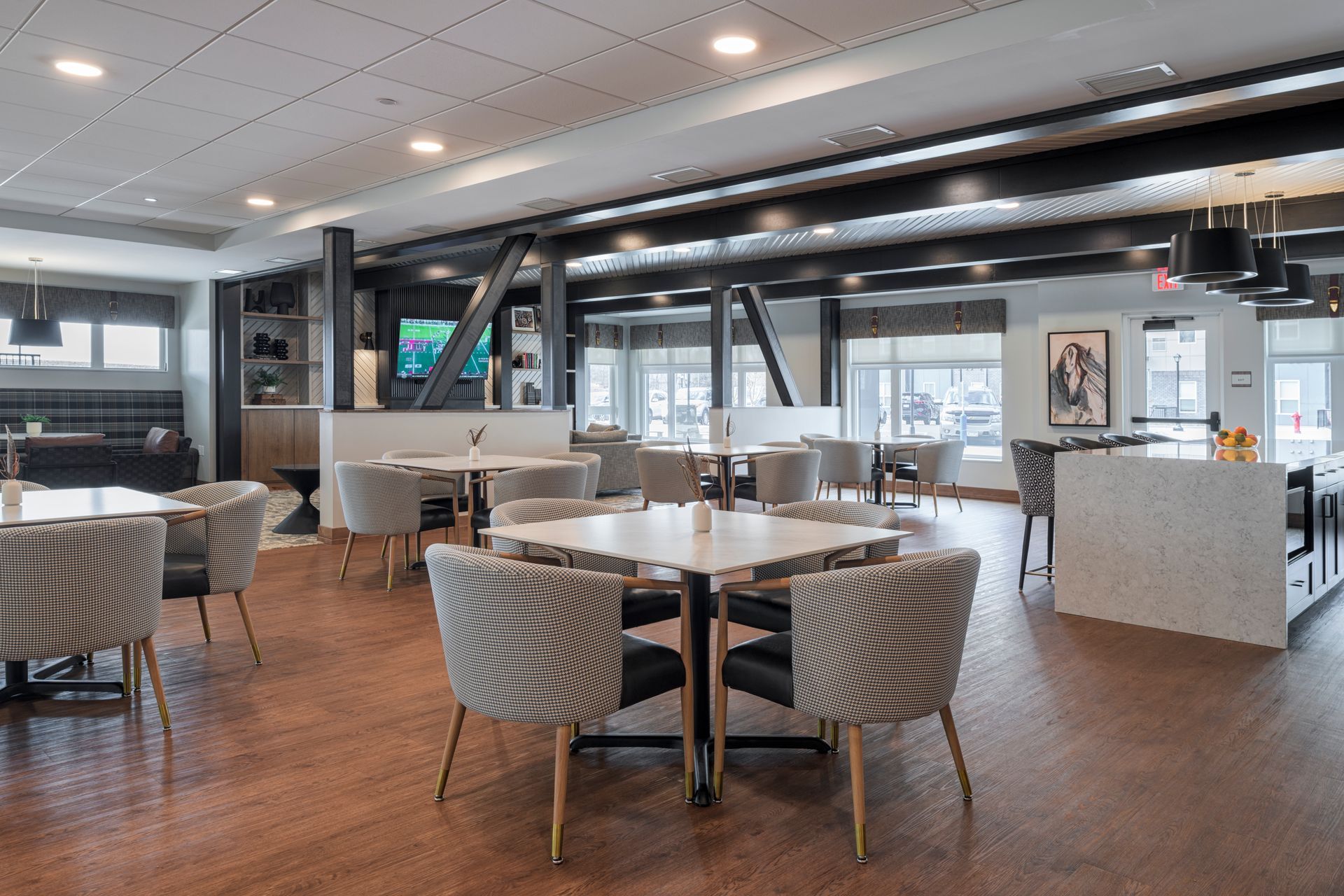 Modern restaurant dining area with light square tables, patterned armchairs, wood flooring, and black structural beams.