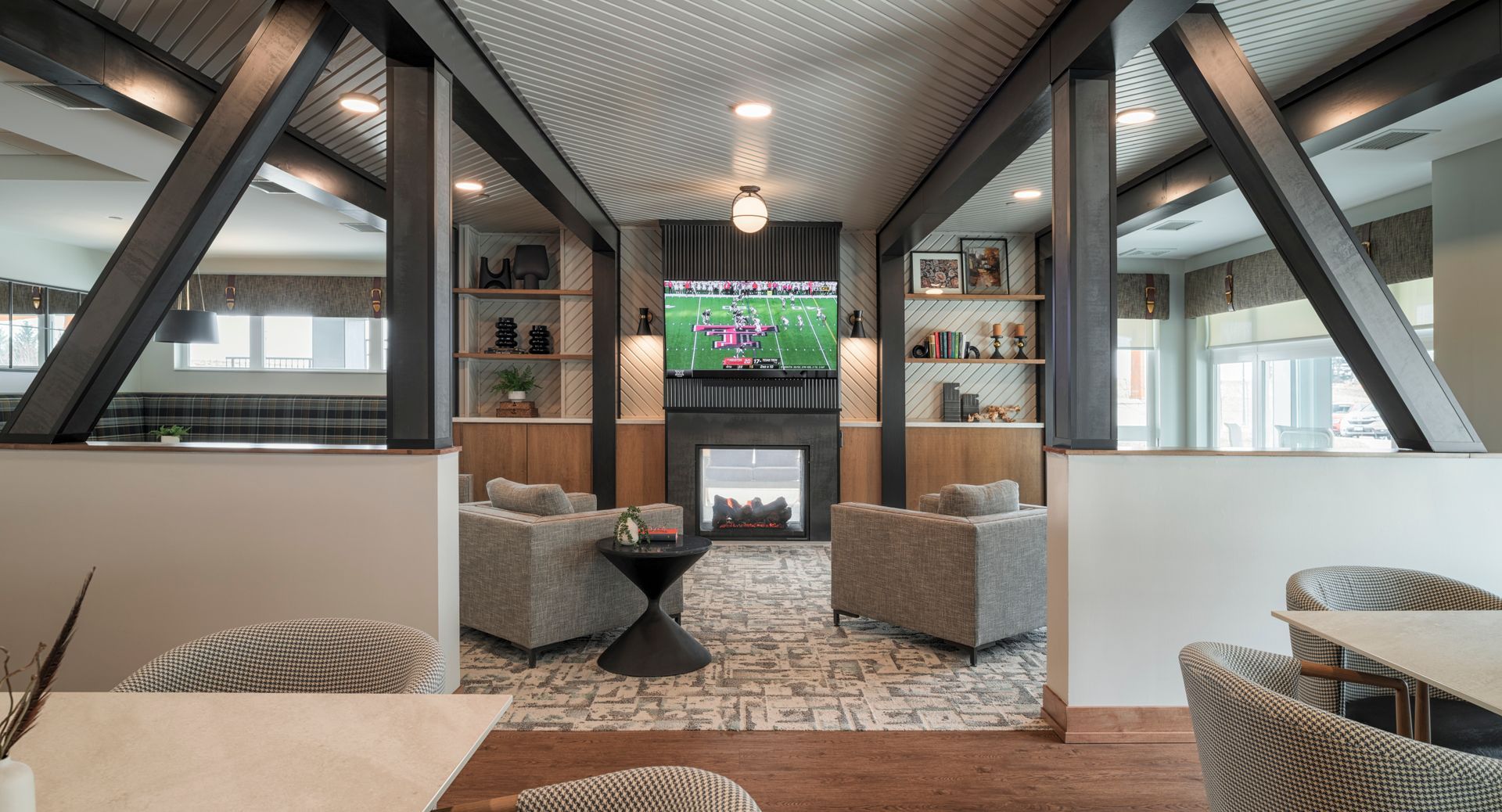 A modern communal lounge featuring a central fireplace, mounted television, two patterned armchairs, and wood accents.