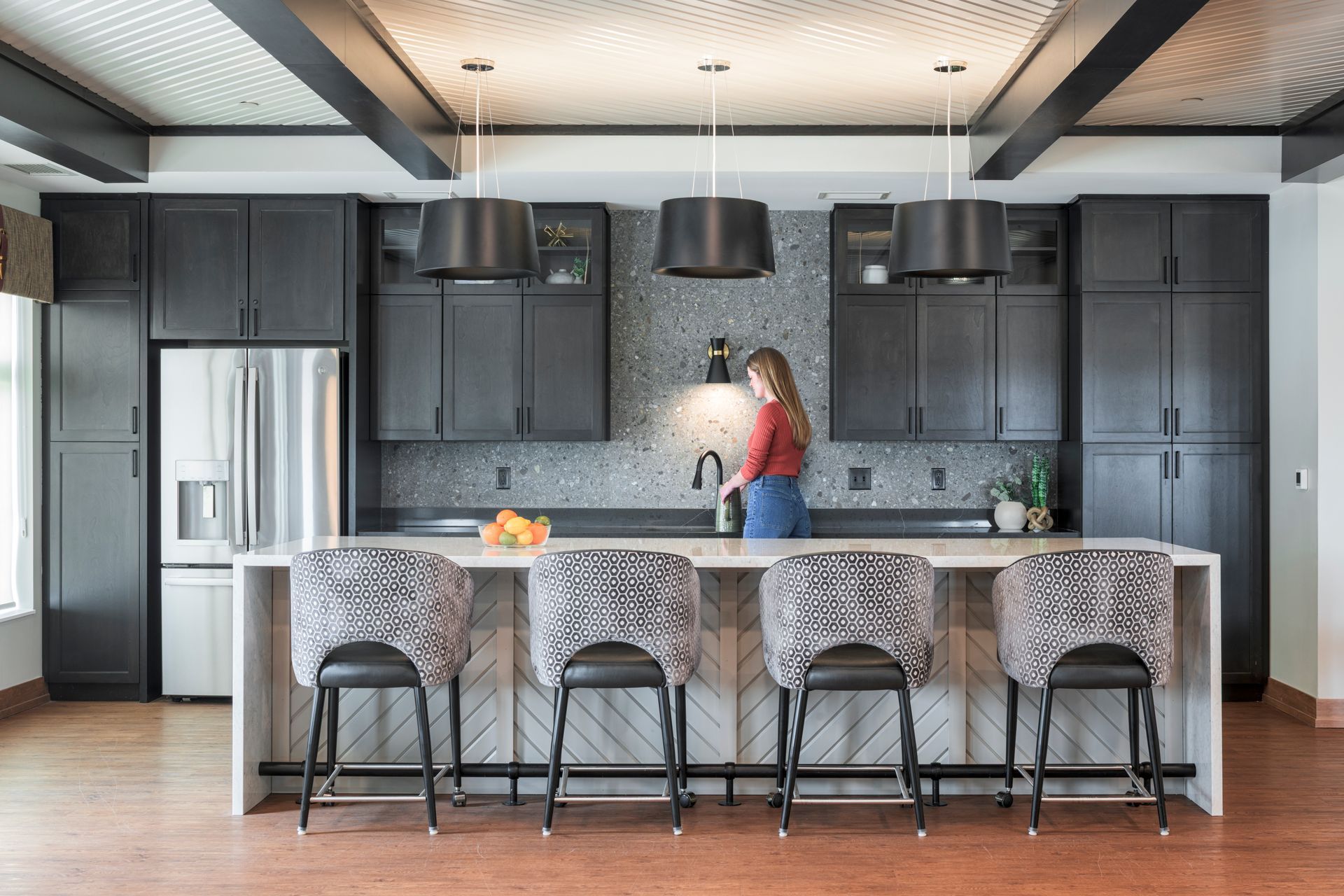 A person stands at a kitchen island in a room with black cabinets, patterned ceiling, and four patterned bar stools.