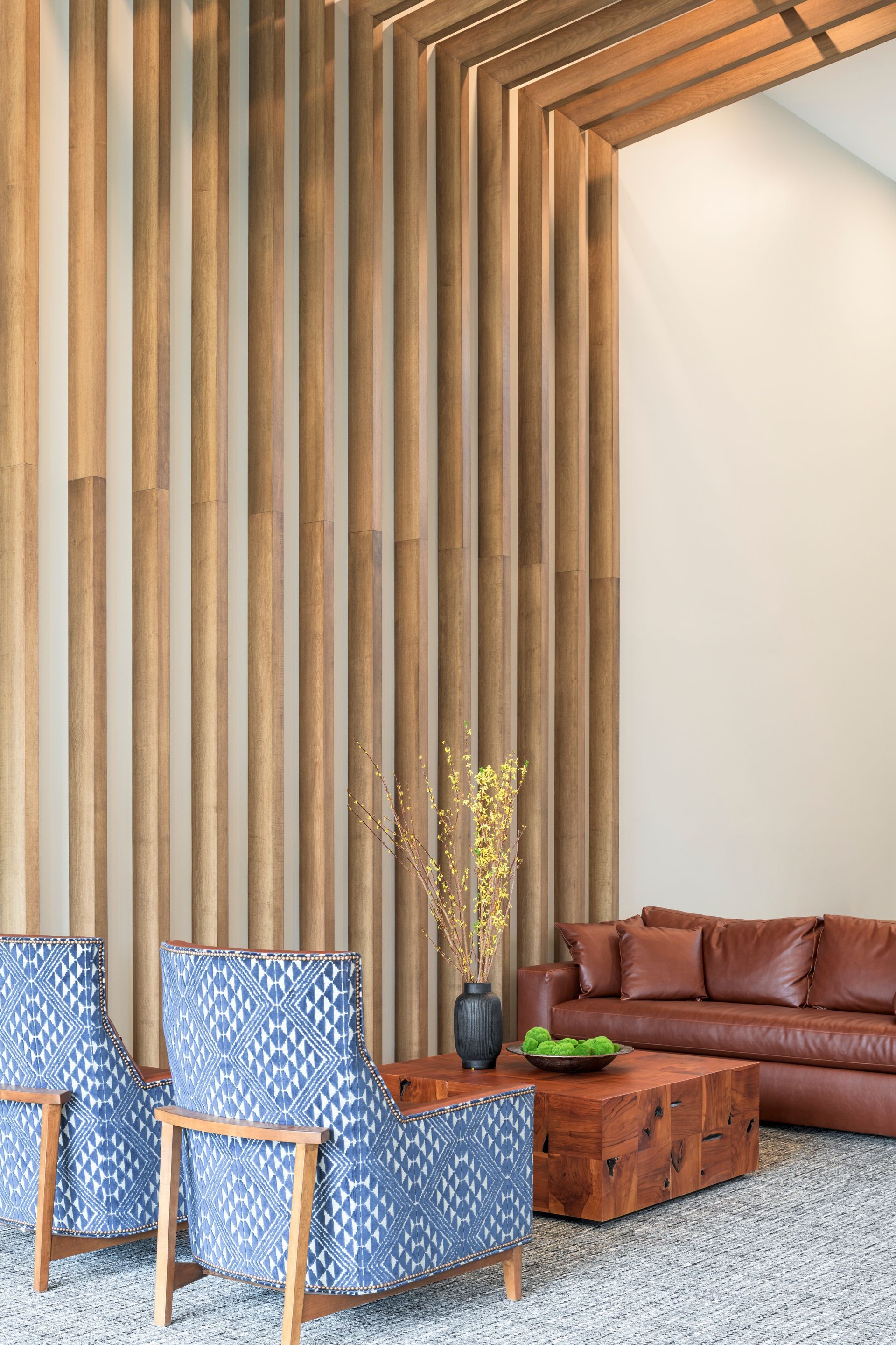 A cozy lounge area featuring two blue patterned armchairs, a leather sofa, and a wooden coffee table against wood slats.