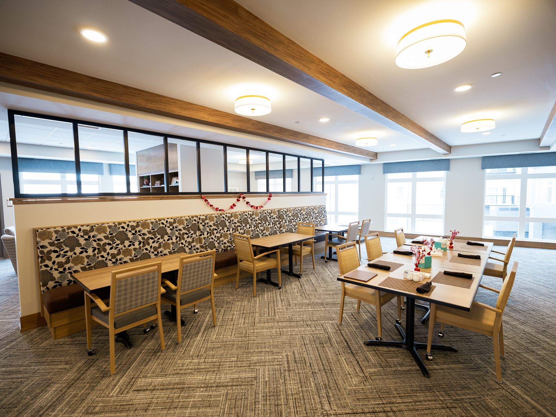 A bright, modern dining area with tables, patterned booth seating, chairs, and large windows.
