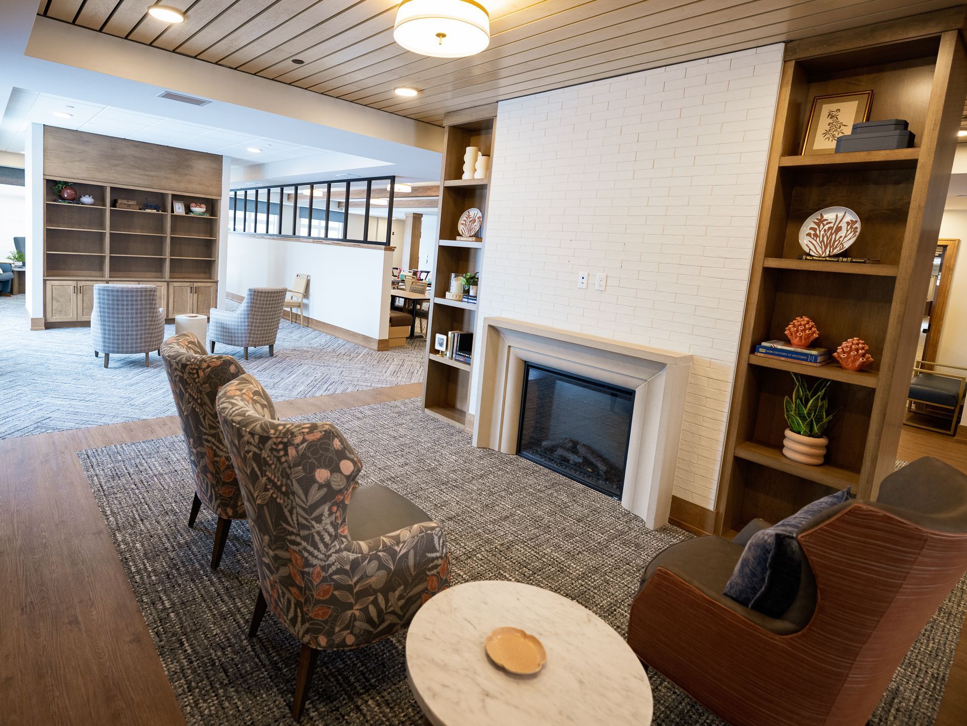 A cozy lounge with floral armchairs, a fireplace, and bookshelves, set on a textured rug in a bright, modern room.