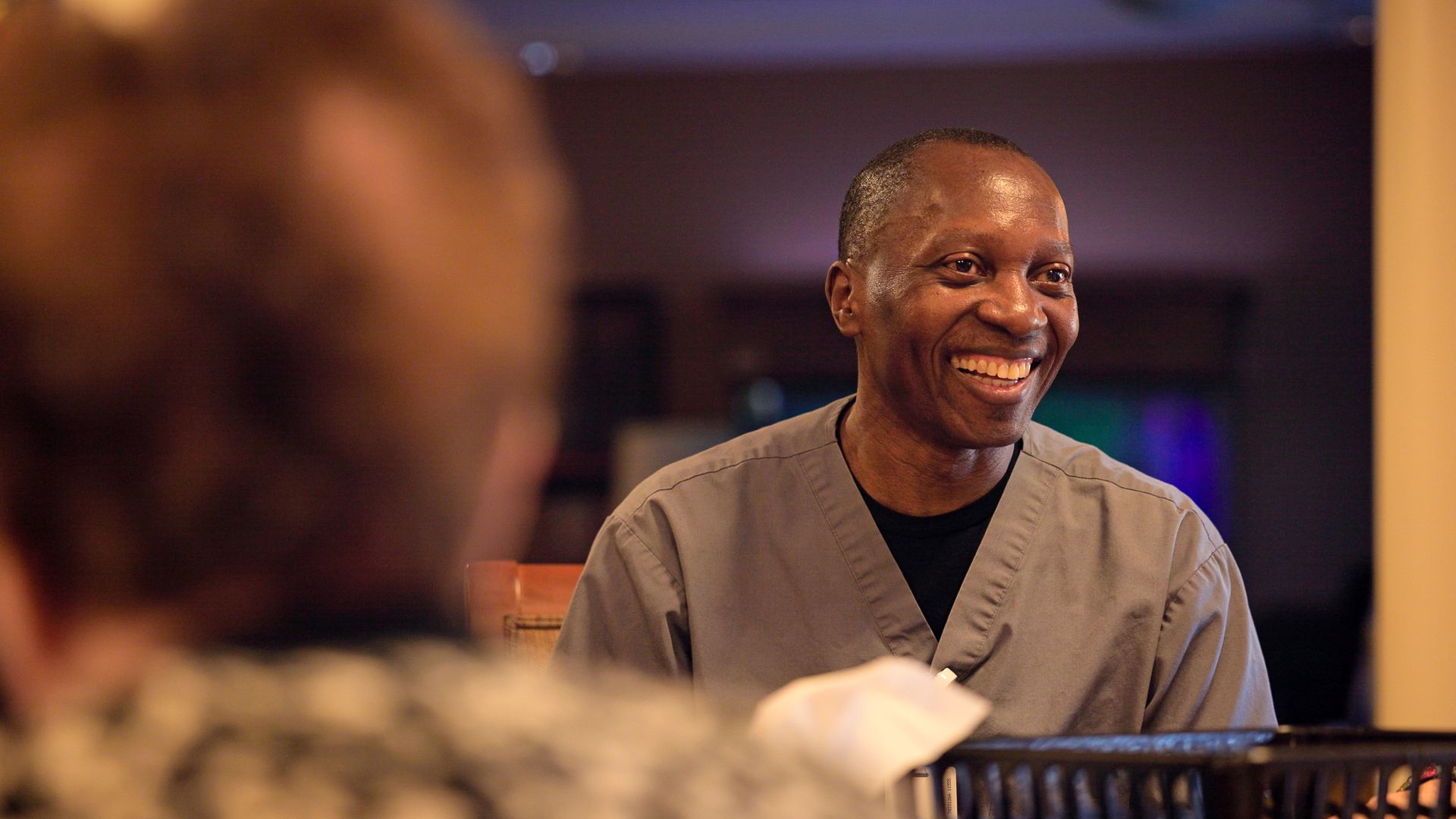 A smiling person wearing grey scrubs looks toward someone out of frame in a dimly lit, soft-focus interior setting.