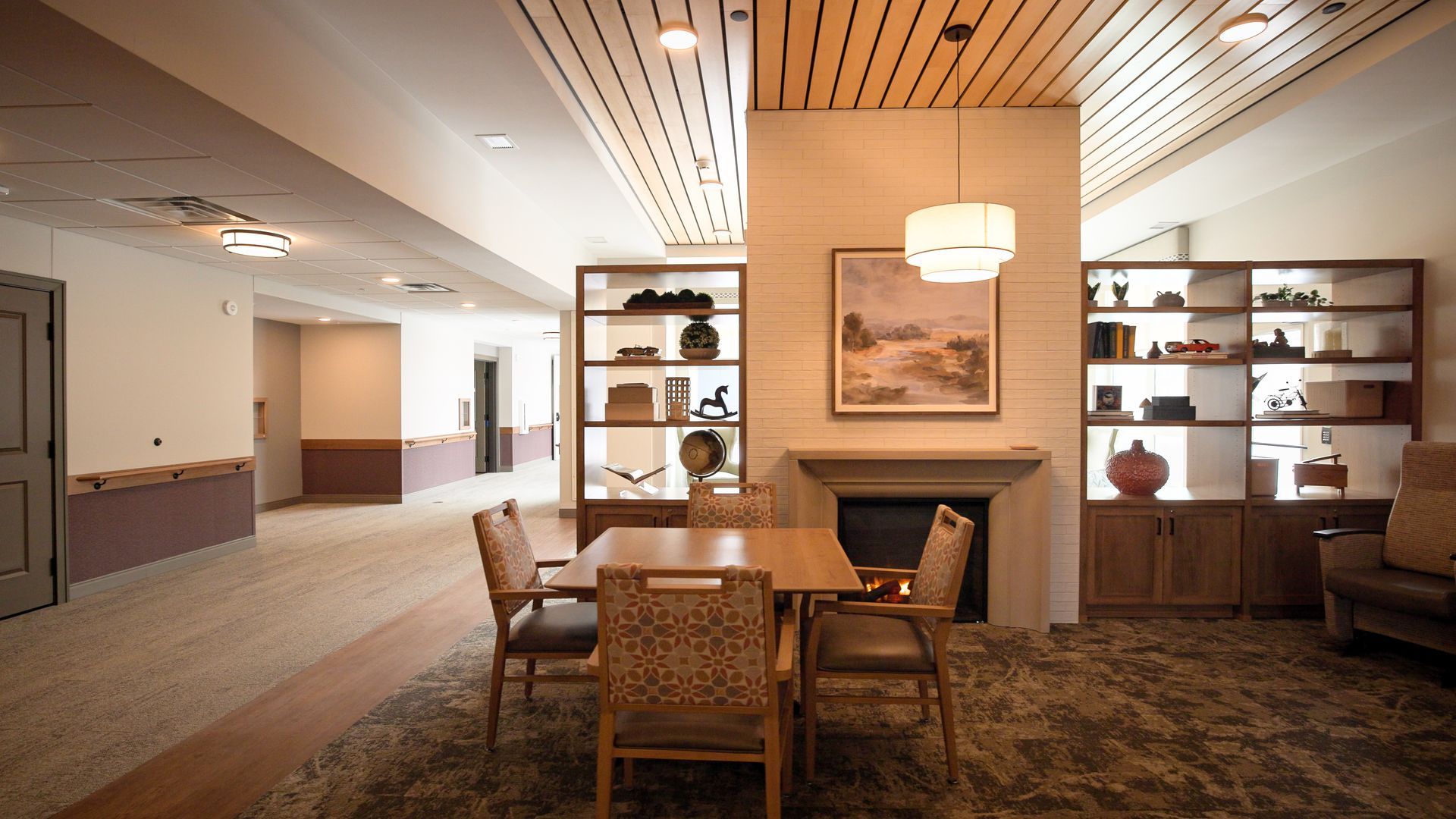 A communal area with a square dining table, chairs, a fireplace, and shelving units under a modern wooden slat ceiling.