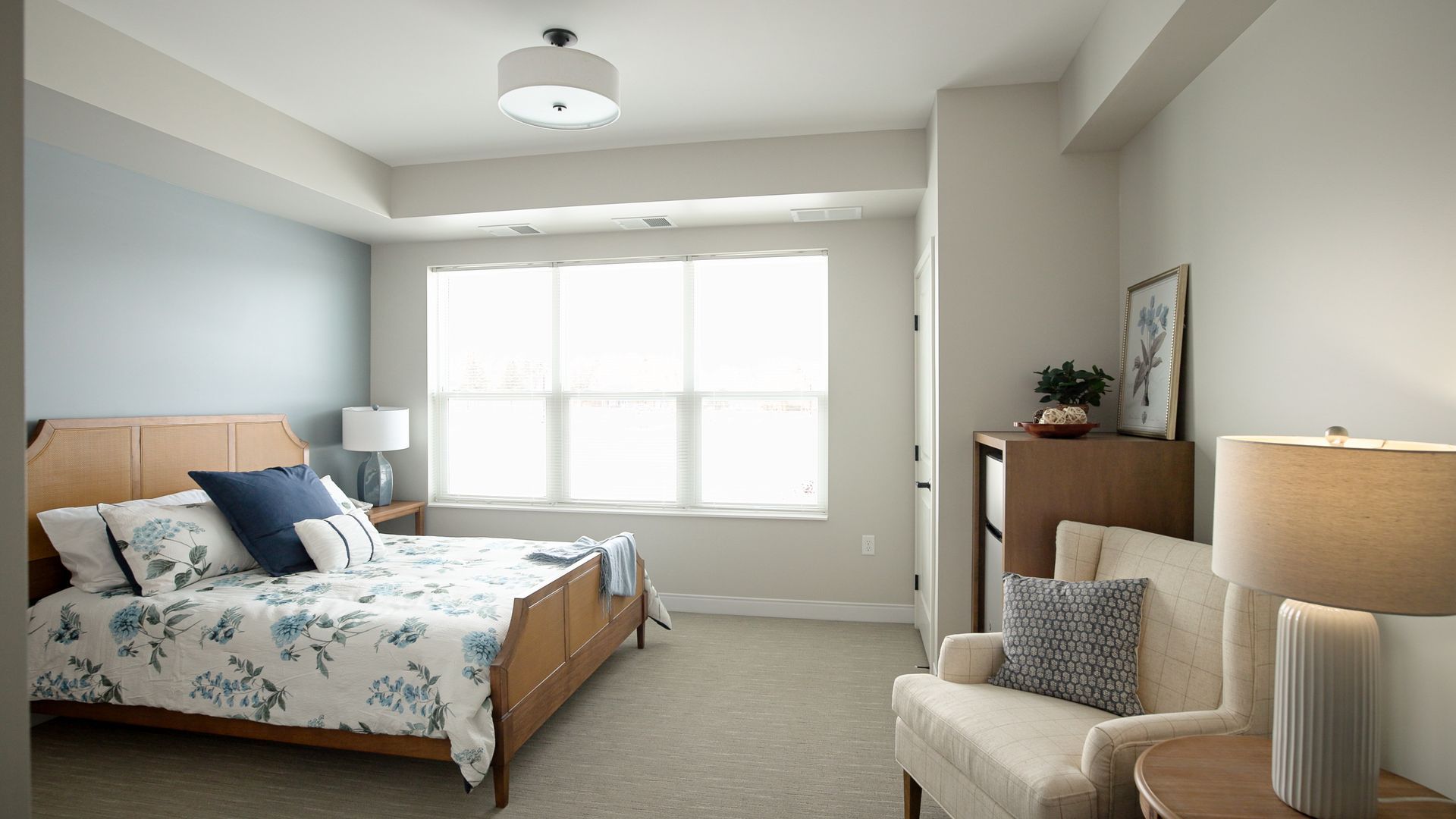 A modern bedroom with a floral bedspread, a blue accent wall, a large window, a beige armchair, and a side table.