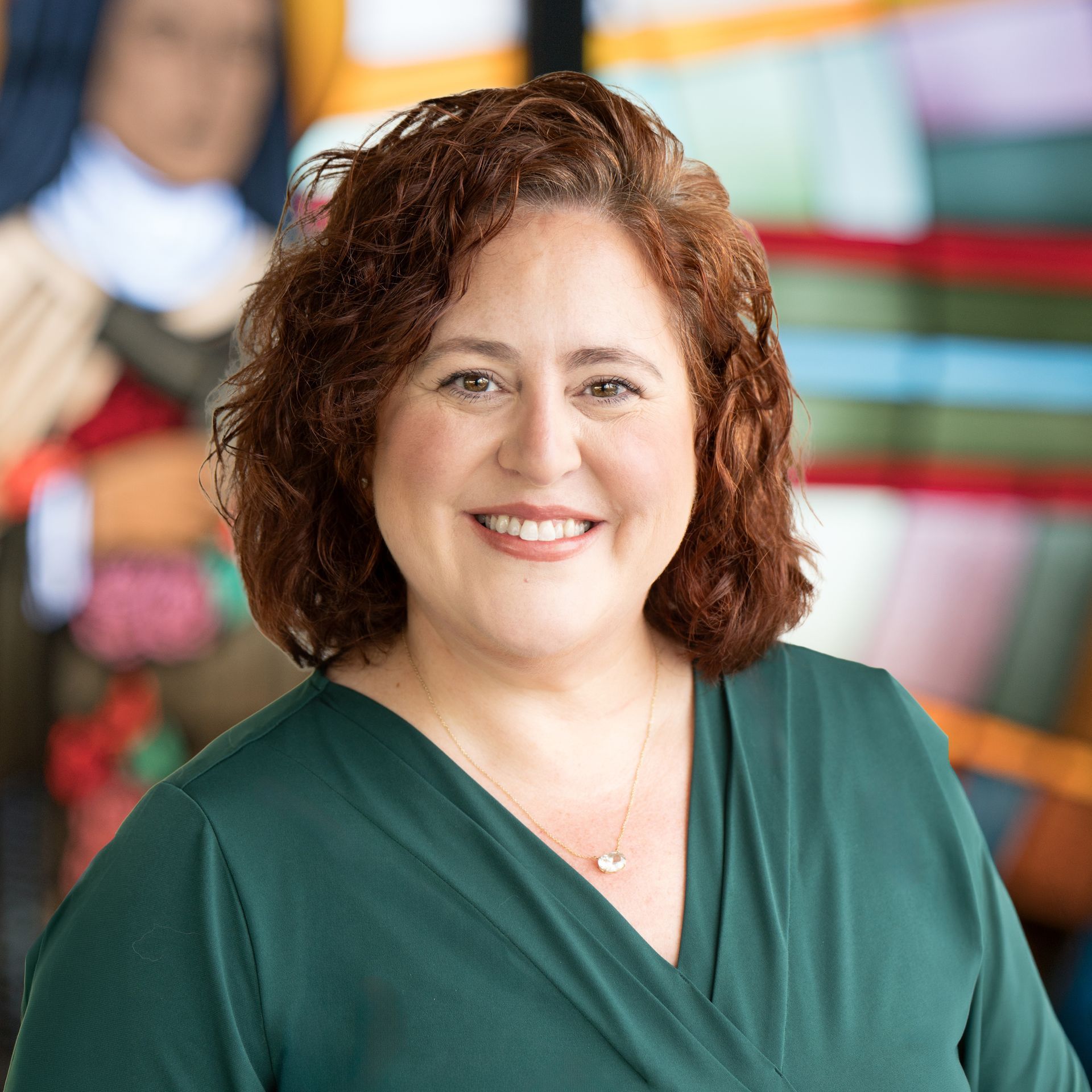 Smiling woman with shoulder-length wavy auburn hair wearing a green V-neck top in front of a colorful backdrop.