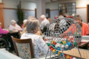 A wire bingo cage filled with colorful numbered balls sits in the foreground, with people playing bingo in a blurry room.