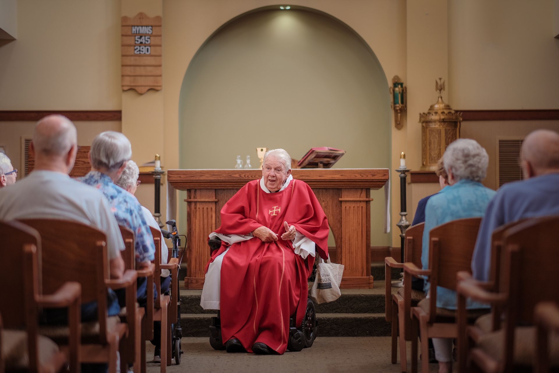 An elderly person in red religious vestments sits in a wheelchair at the front of a church, addressing an audience.