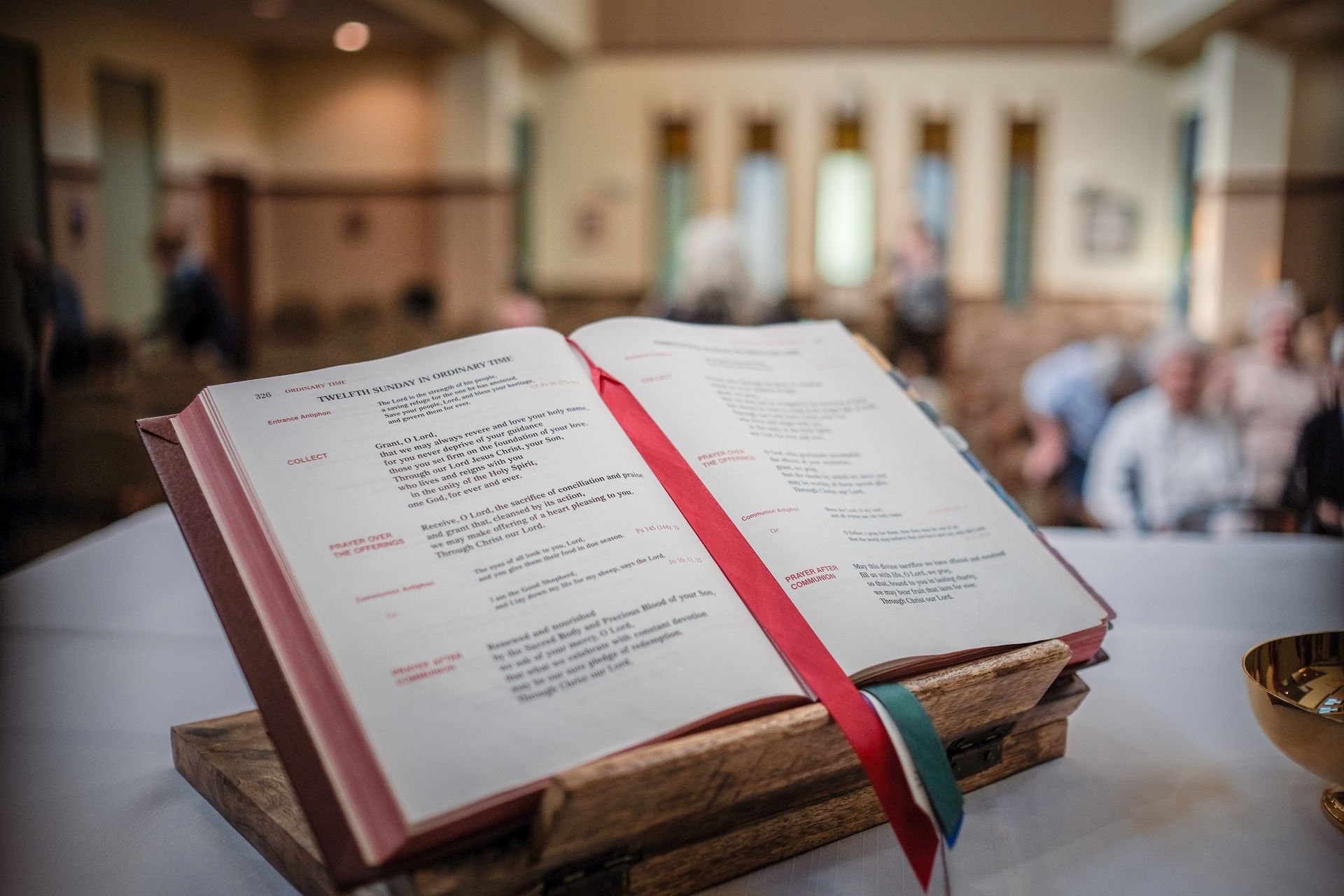 An open liturgical book sits on a wooden stand on a table, with a blurred church congregation in the background.