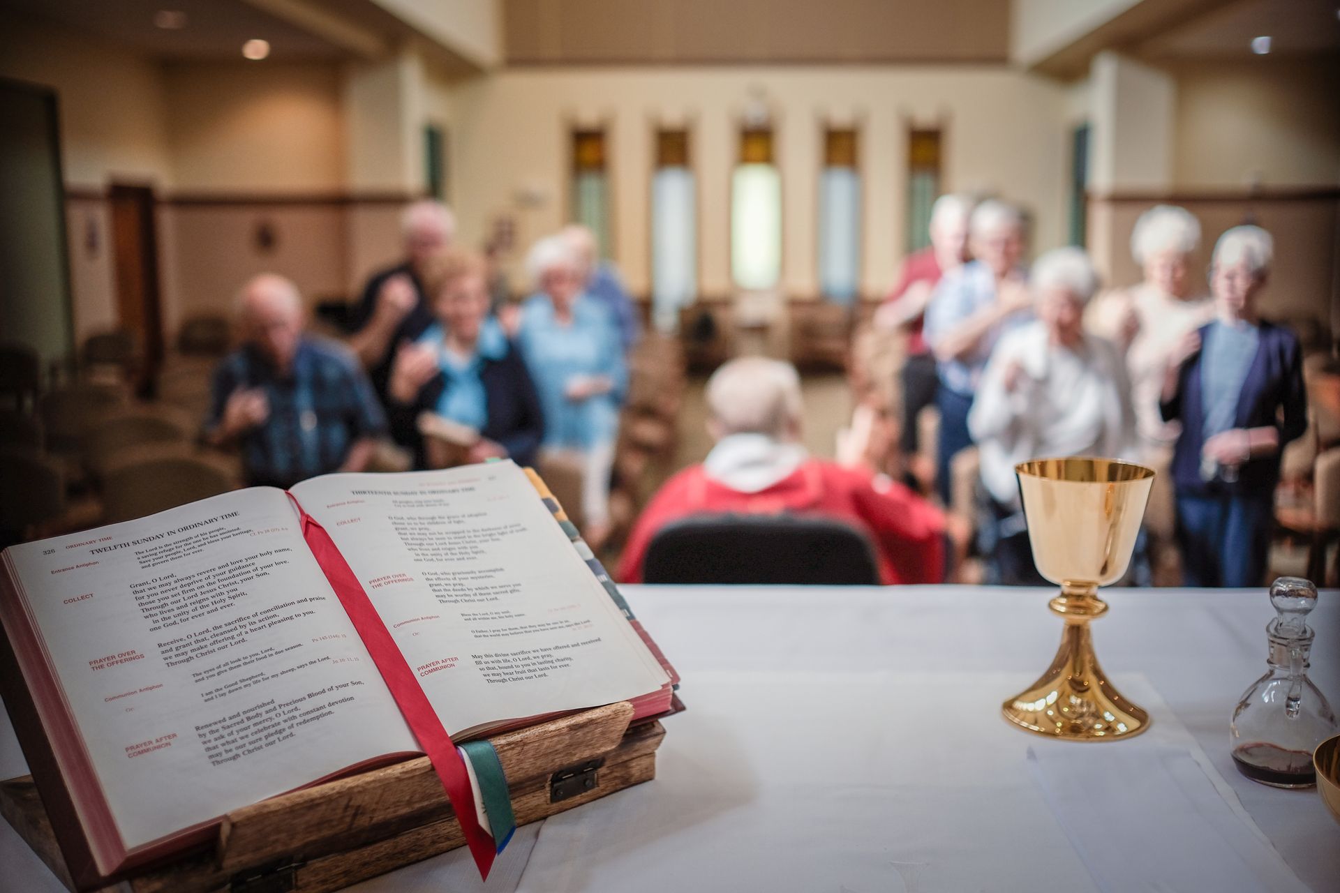 A book and golden chalice sit on a white altar with a blurred group of people gathered in a church background.