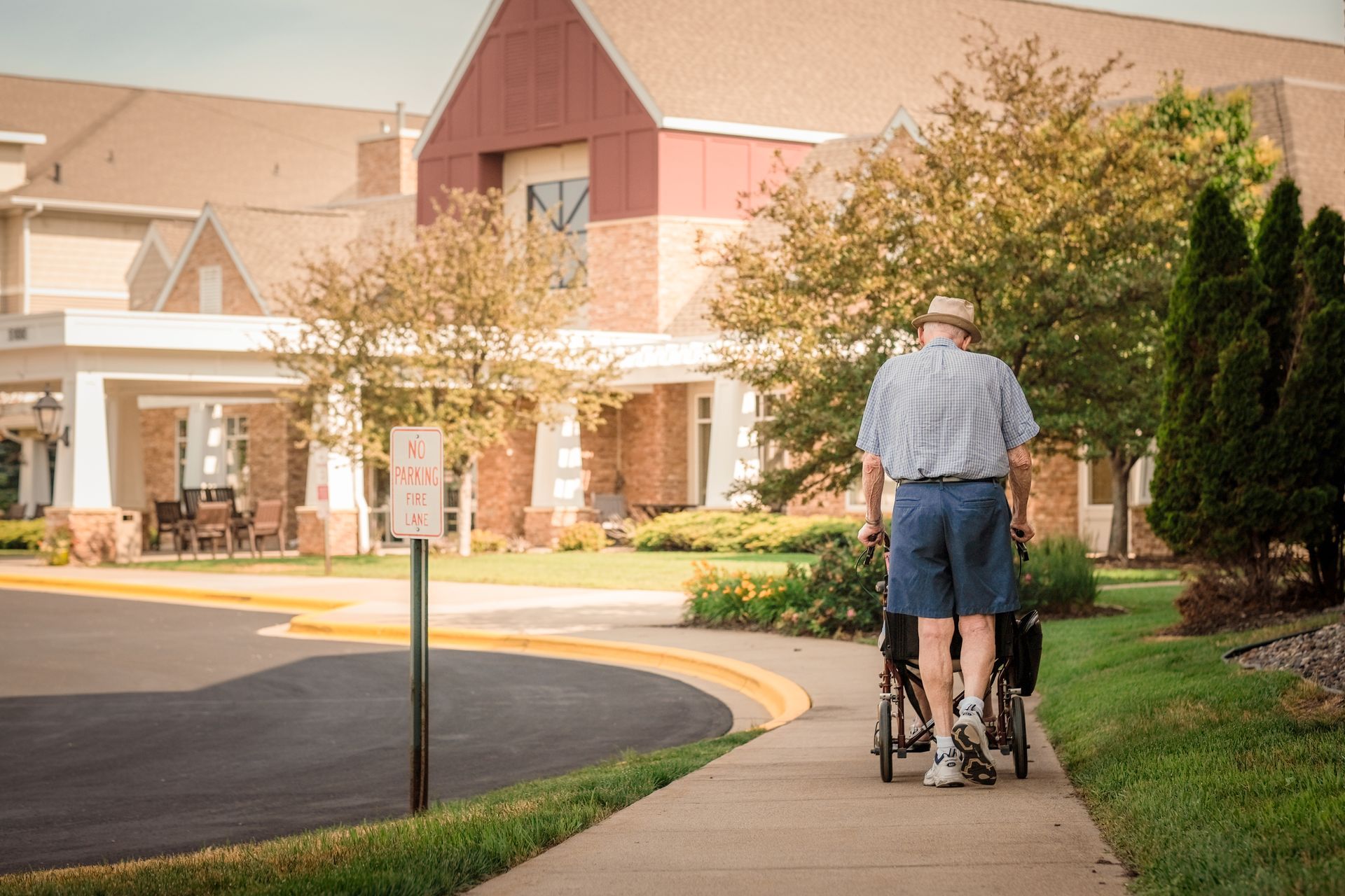 A person wearing a hat and shorts walks on a sidewalk while pushing a wheelchair toward a large building.