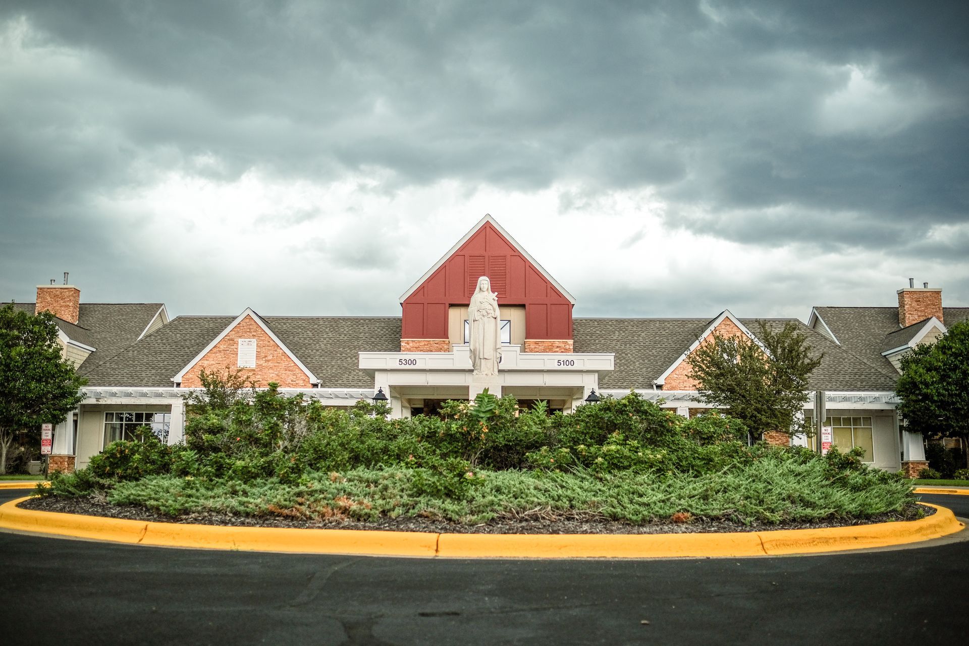 A statue stands in a circular garden bed in front of a sprawling, multi-gabled building under a cloudy sky.