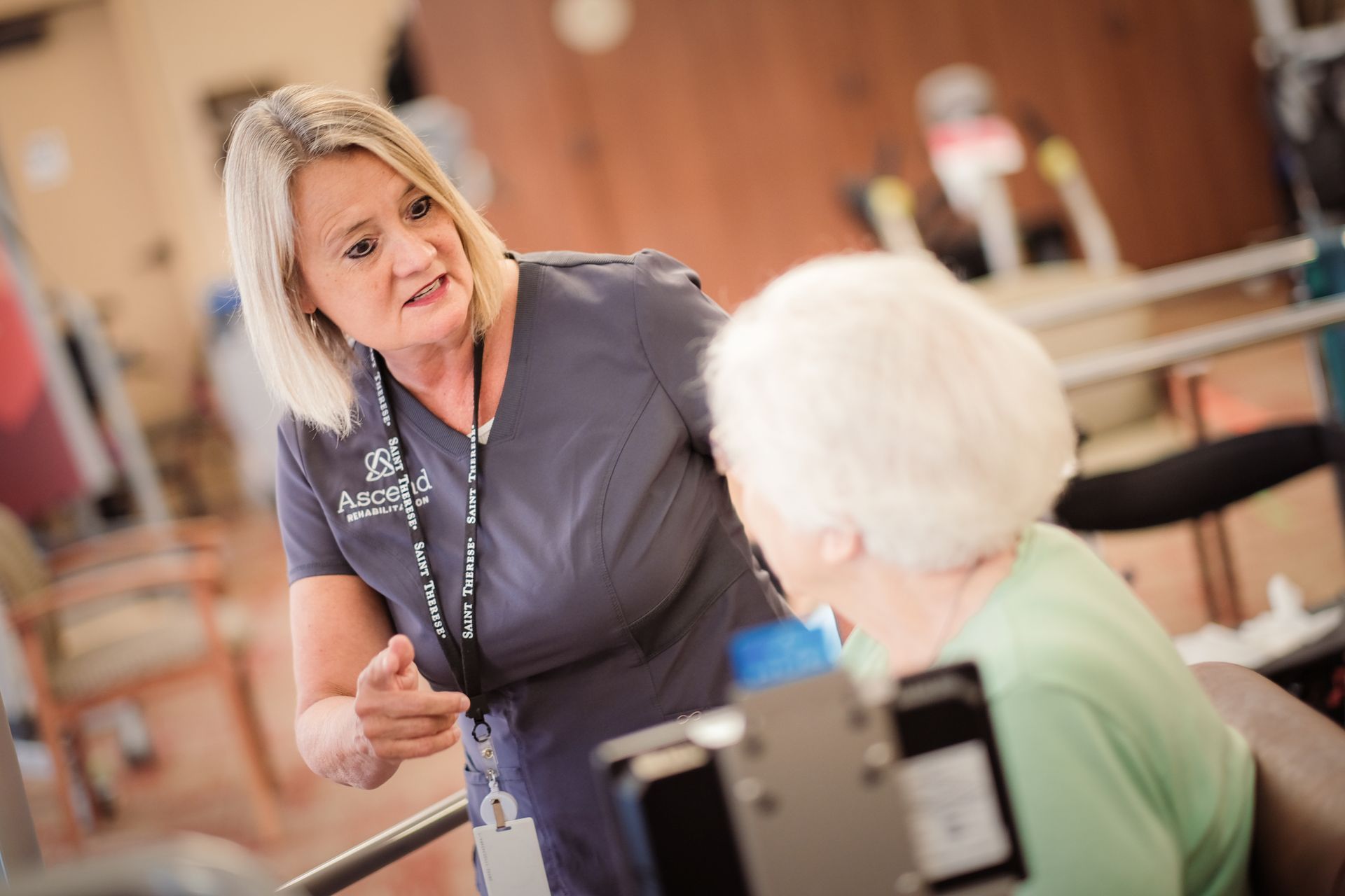 A healthcare professional in a grey uniform speaks with a person in a medical setting, pointing towards equipment.