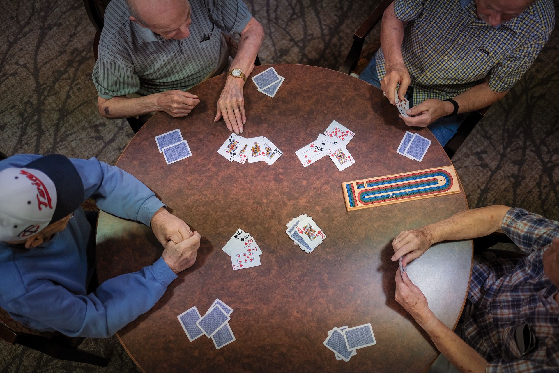 Four people play a game of cribbage around a circular table, with cards laid out and a wooden scoring board in the center.