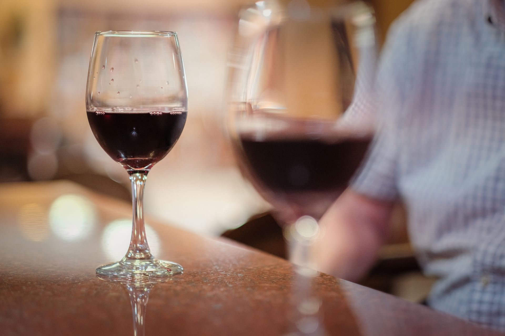 Two glasses of red wine sit on a polished surface, with a person blurred in the background.