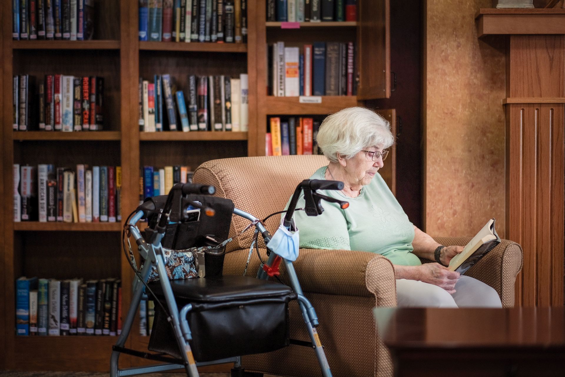 An individual sits in an armchair reading a book next to a walker, with bookshelves in the background.
