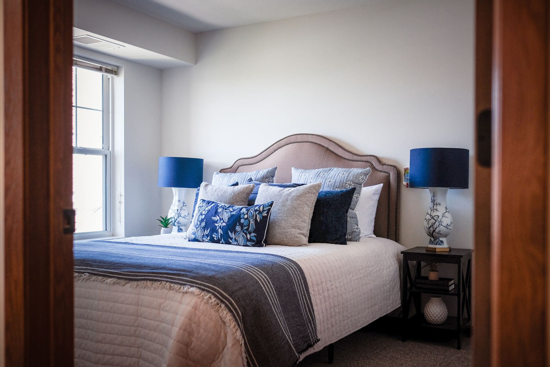 A bedroom with a bed featuring a beige upholstered headboard, blue floral pillows, and a dark blue throw blanket.