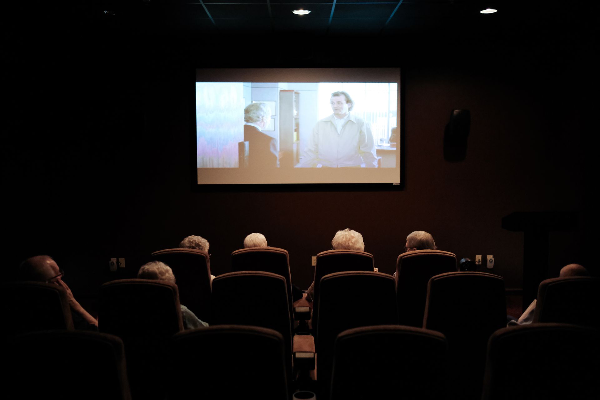 A dimly lit movie theater showing a black-and-white film to a small audience seated in dark, high-backed chairs.