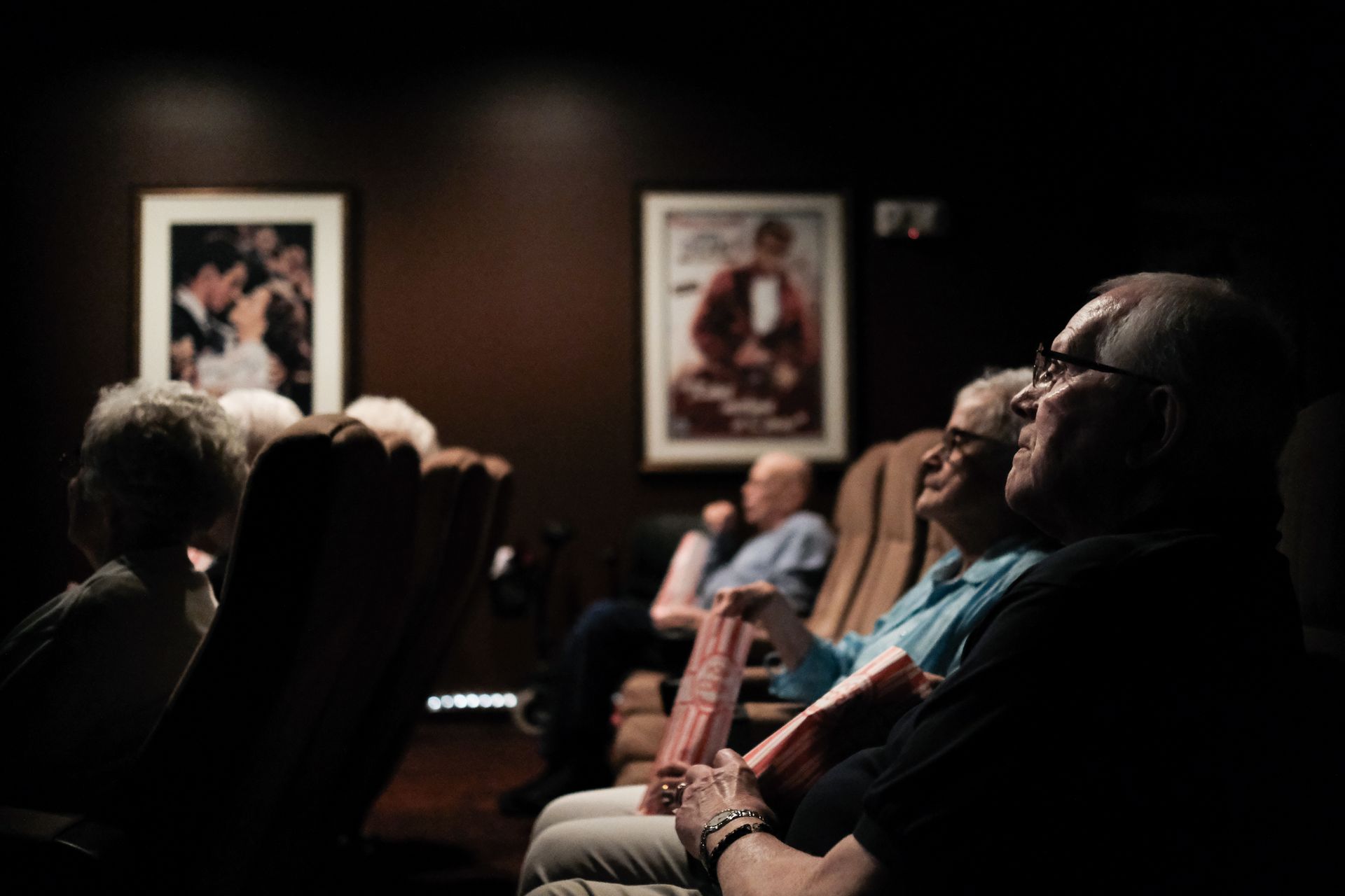 People sit in a dimly lit cinema, watching a screen with framed vintage movie posters on the wall behind them.