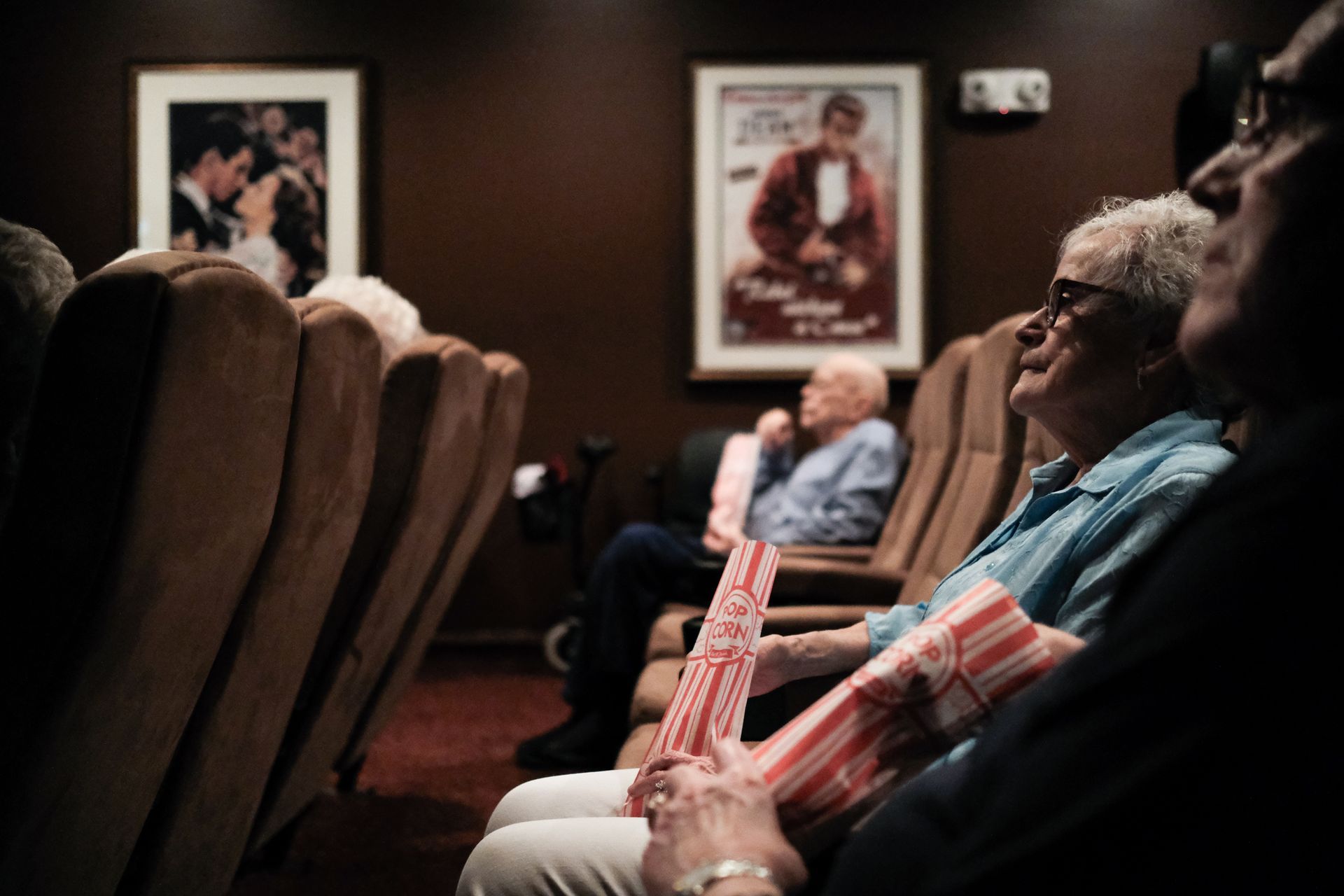 People sit in a dimly lit theater watching a movie, holding boxes of popcorn, with vintage movie posters on the walls.