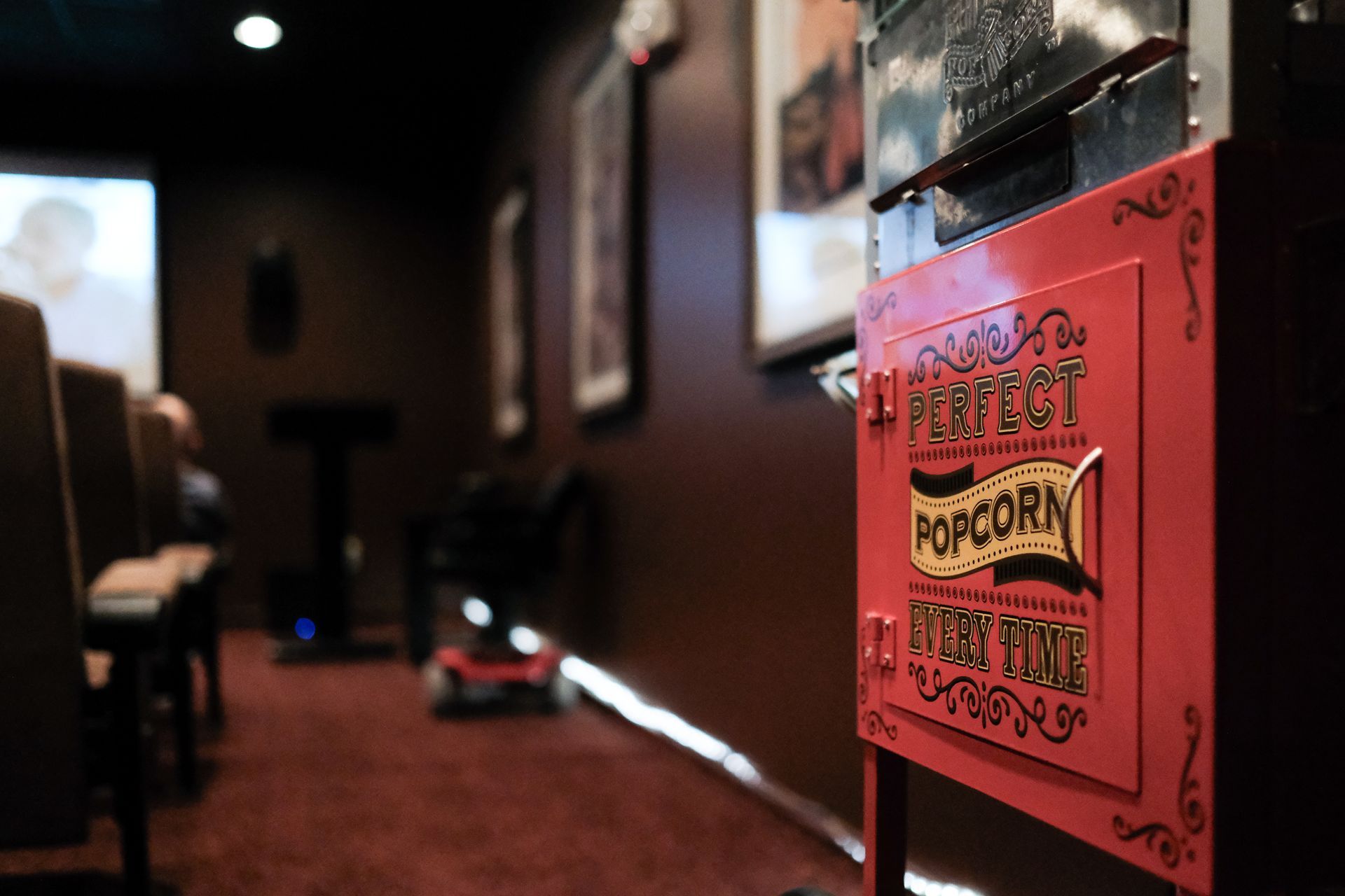 A red popcorn machine in the foreground of a dim movie theater, with rows of seats and a screen visible in the distance.
