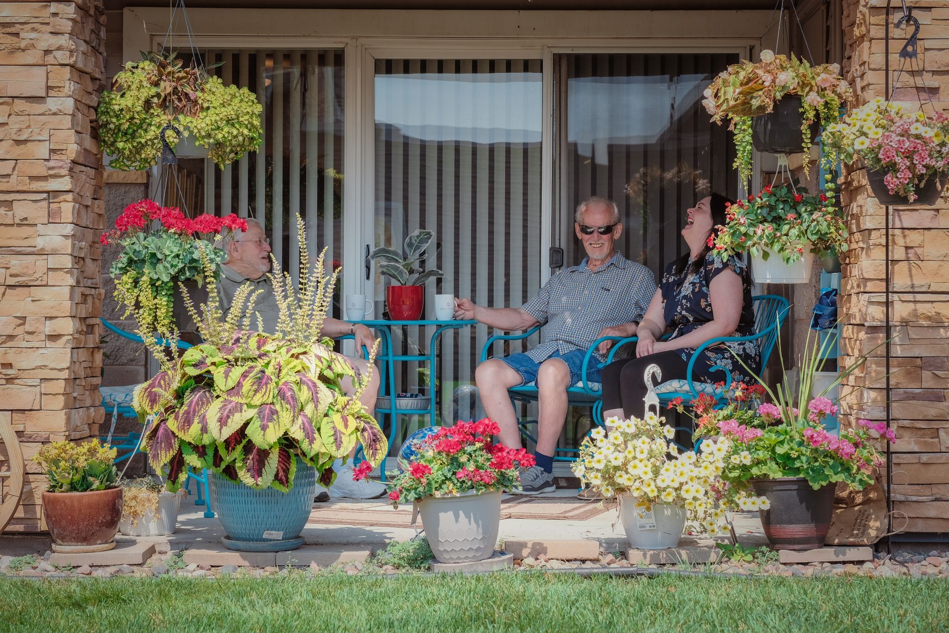 Three people relax on a patio surrounded by many colorful potted plants and hanging flowers.