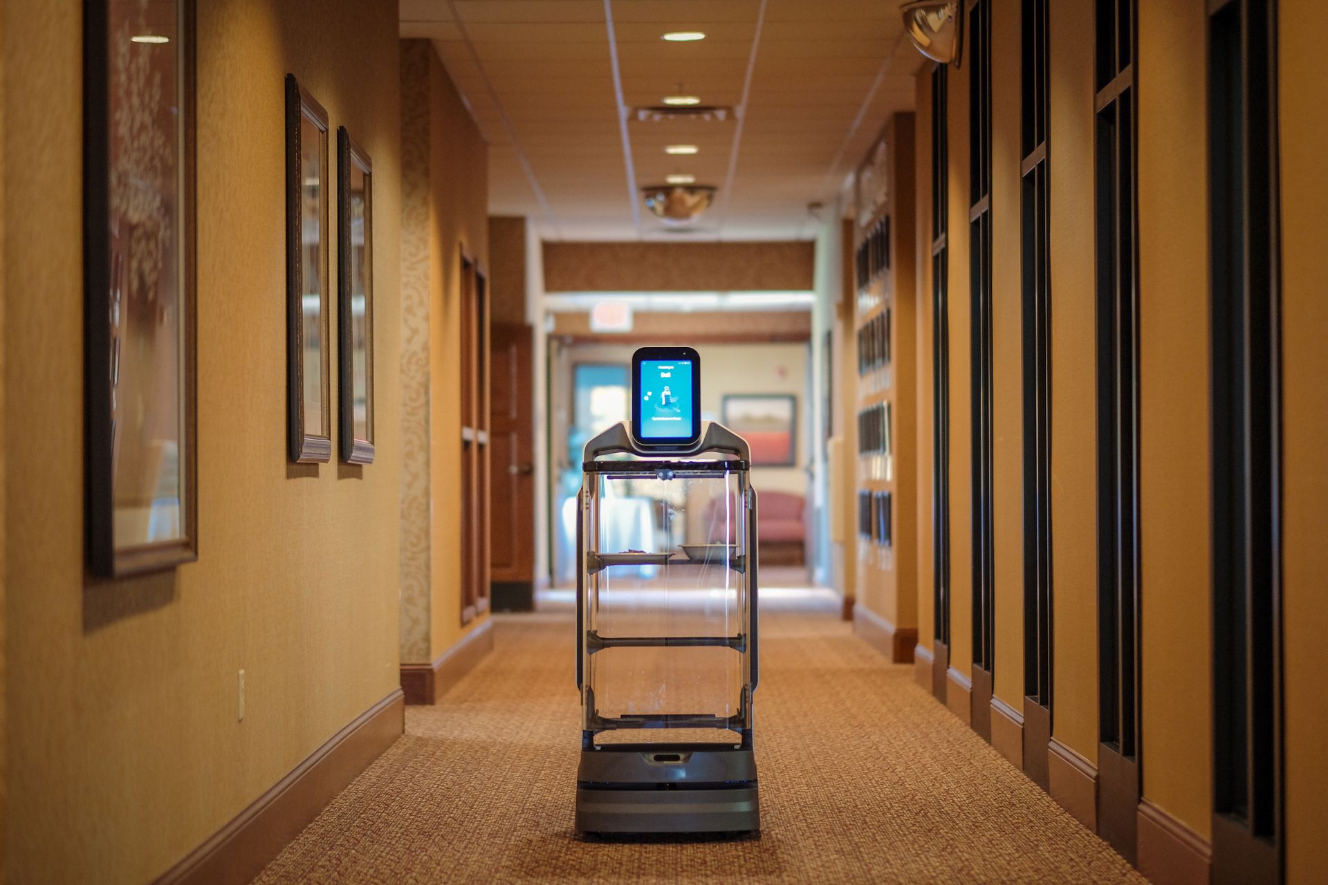 A robotic delivery cart with a glowing digital display sits in a carpeted, indoor hallway.
