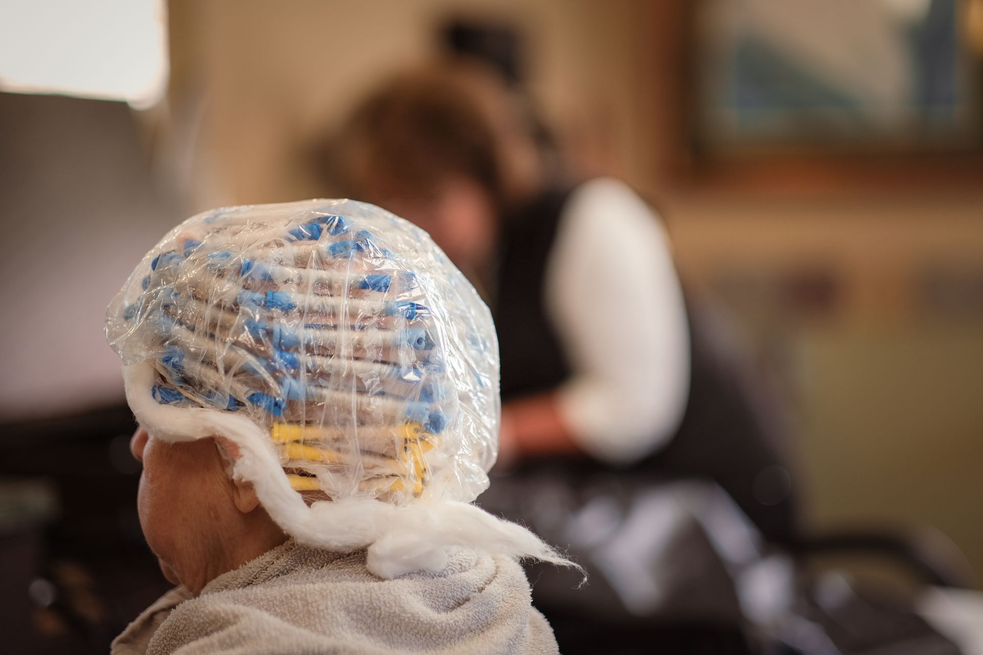 A person wears hair rollers covered by a plastic processing cap while sitting in a salon chair.