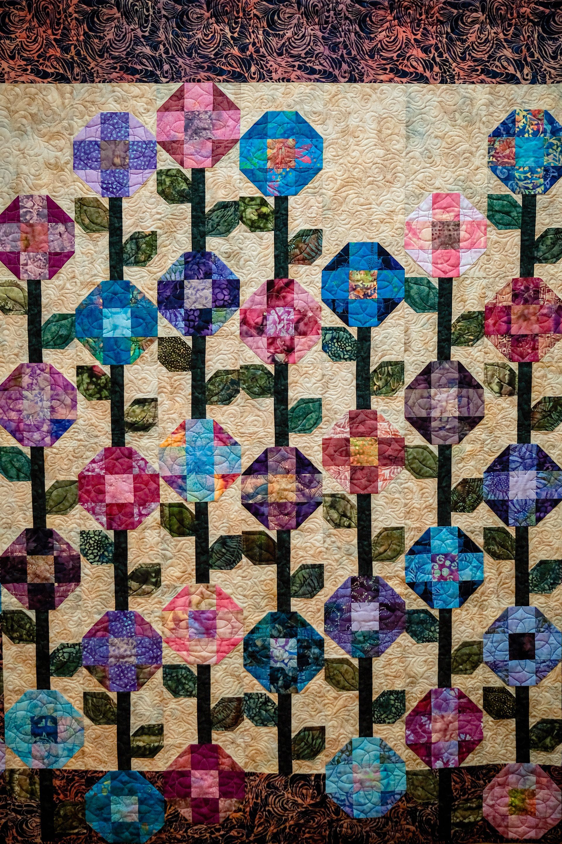 A quilt featuring vertical rows of colorful, patchwork-style flowers in various shades of blue, pink, and purple.
