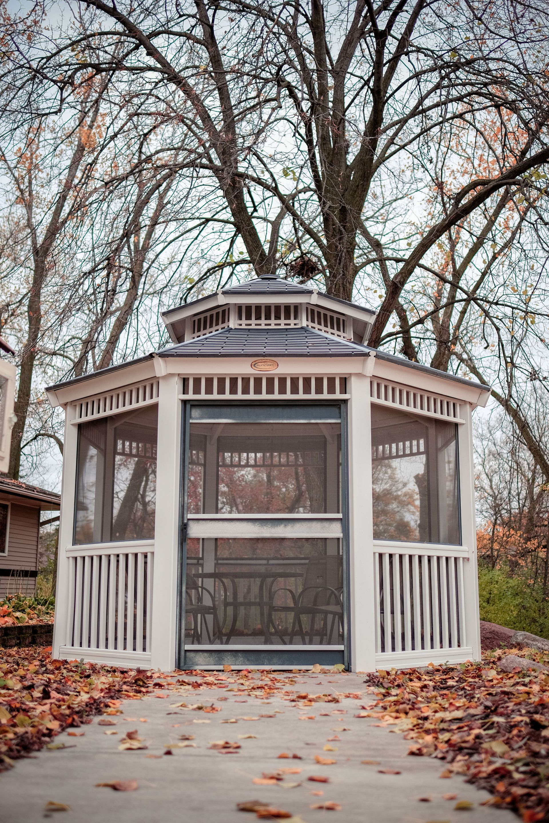 A white, screened-in wooden gazebo stands on a stone path surrounded by fallen autumn leaves and bare trees.