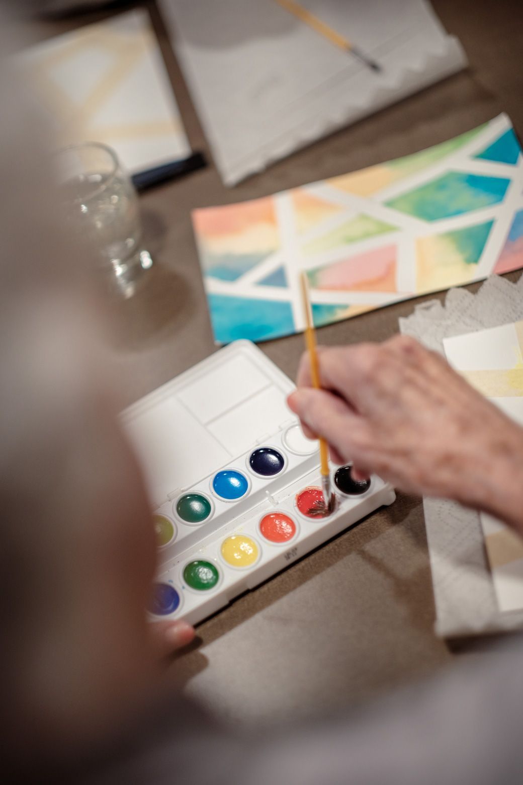 A person uses a brush to pick up red paint from a watercolor palette next to paper with geometric shapes.
