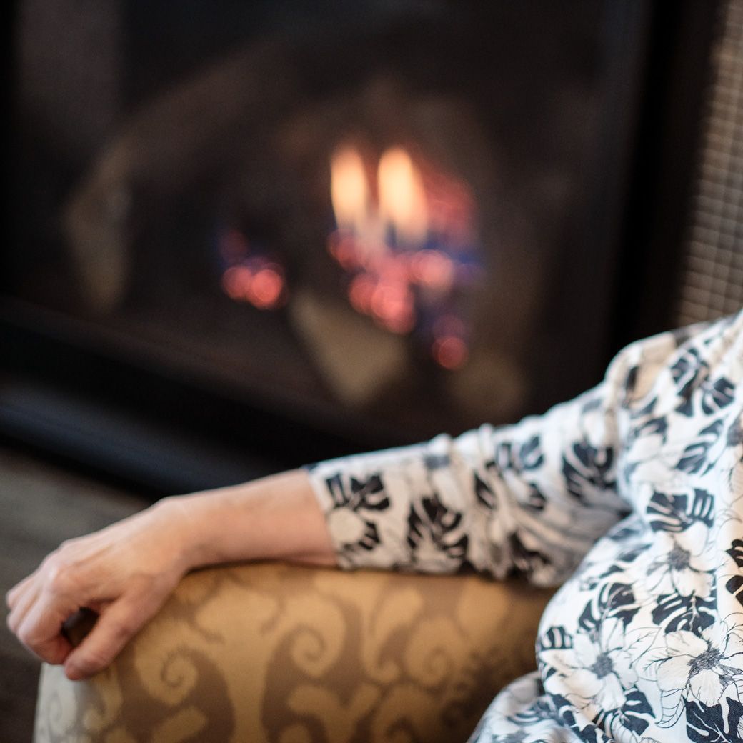 A person wearing a floral print long-sleeve shirt rests their hand on a chair in front of a glowing fireplace.