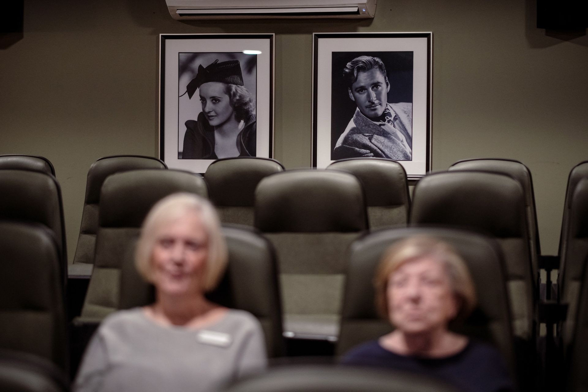 Two individuals sit in a movie theater with framed black-and-white portraits of actors hanging on the wall behind them.