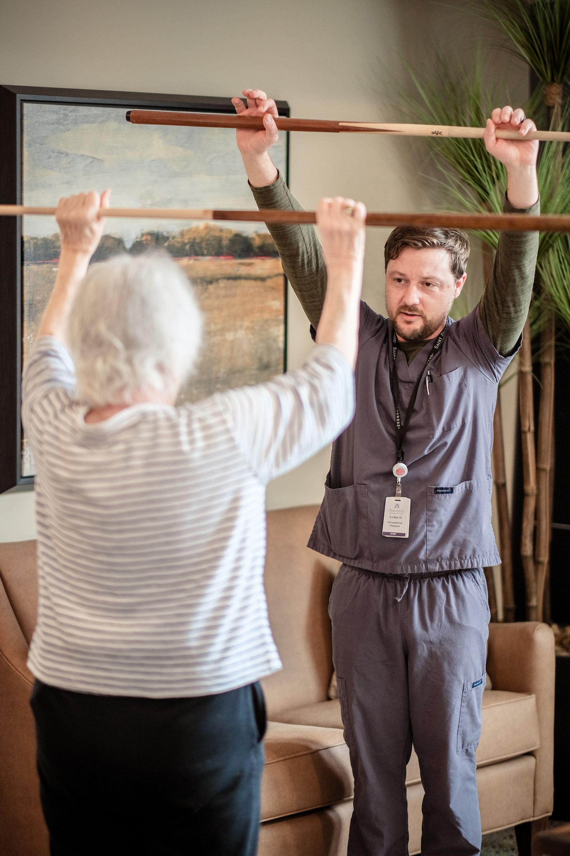 A healthcare worker guides a patient in a physical therapy exercise, both holding a wooden staff raised above their heads.