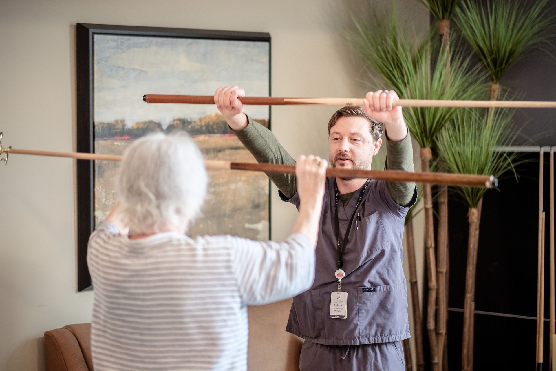 A therapist and a patient perform physical therapy exercises, both holding horizontal wooden rods at chest level.