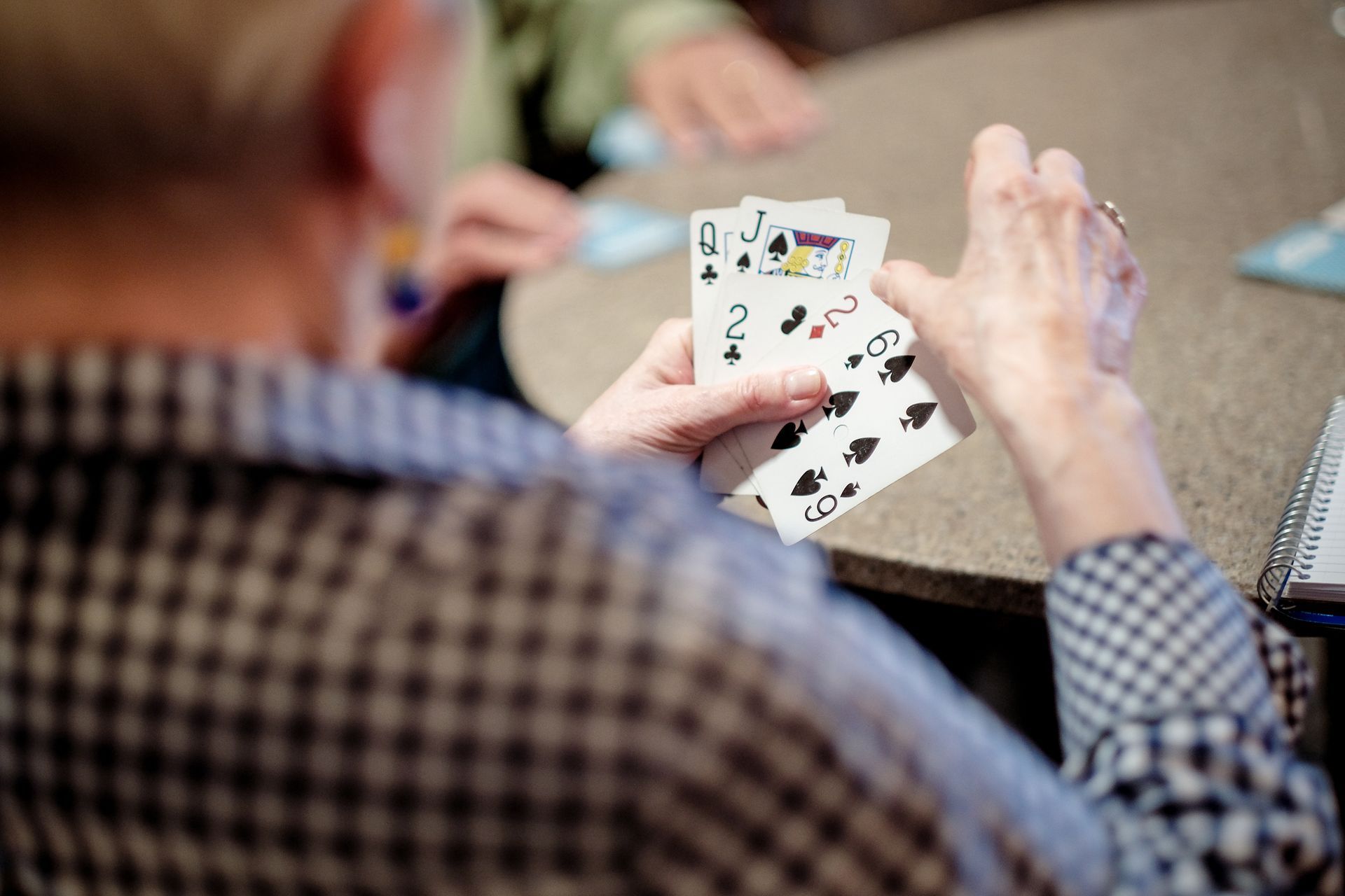 A person holds a hand of playing cards, including a queen of spades, during a card game at a table.