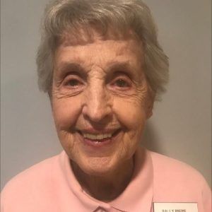 A smiling person with gray hair wearing a light pink collared shirt and a name tag against a plain gray background.