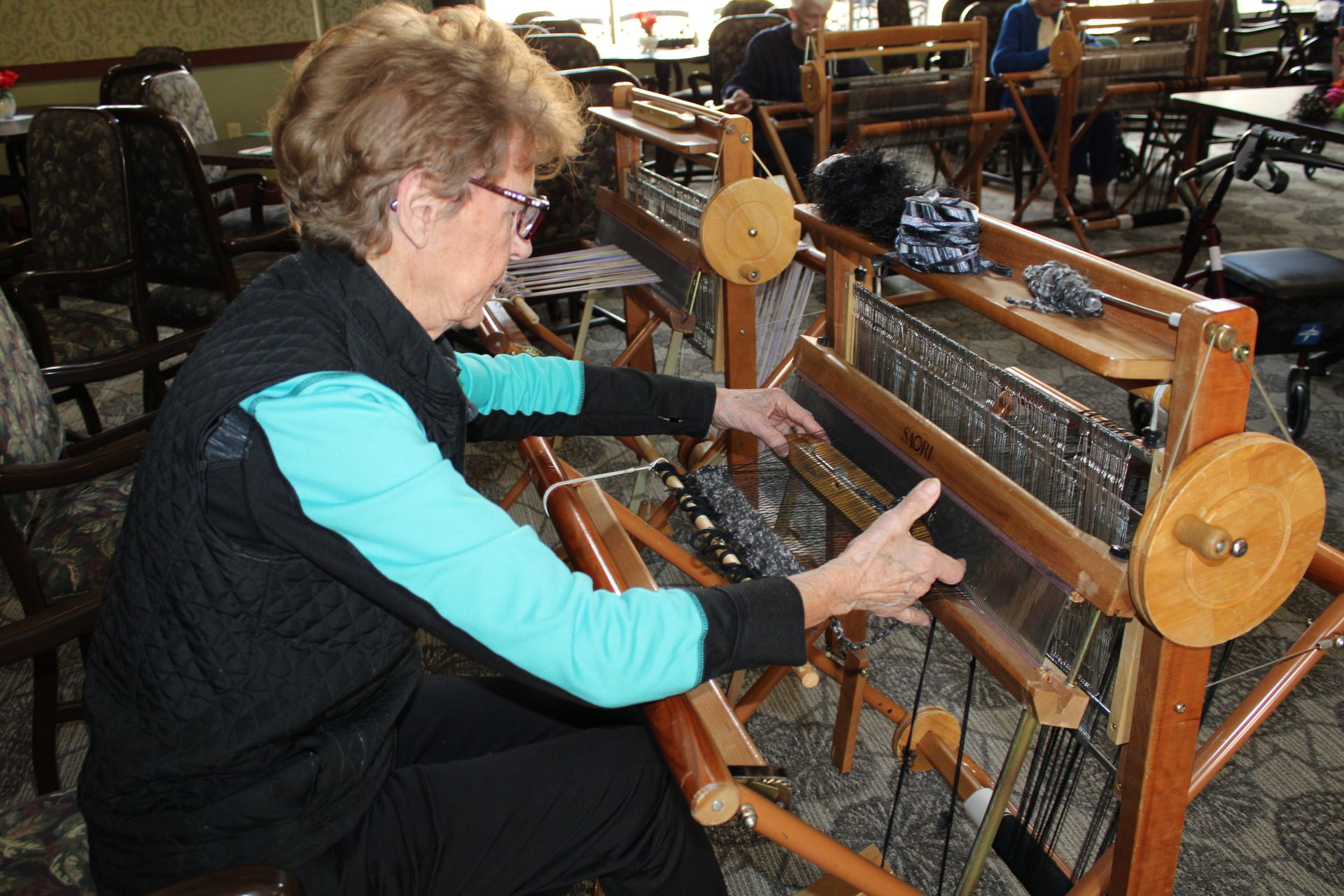 A person weaving with a wooden loom in a communal activity room.
