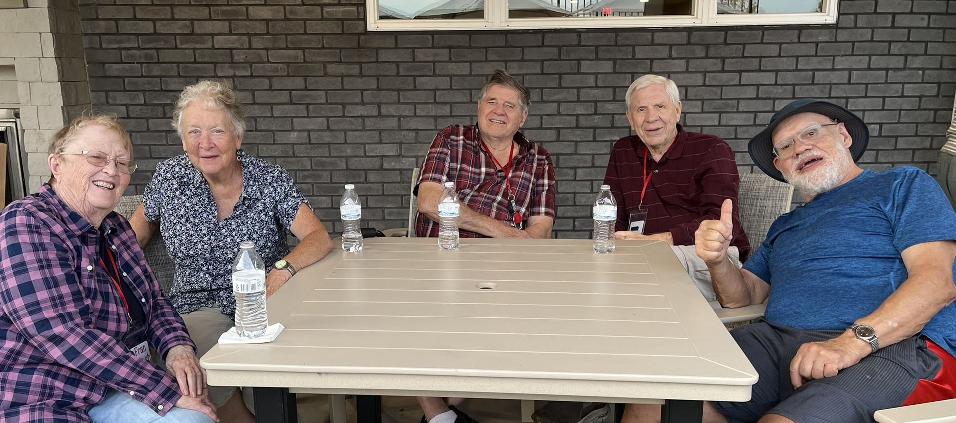 Five people sit at a table on a patio with a brick wall background, smiling and talking.