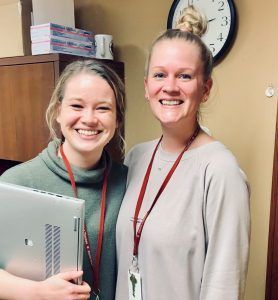 Two people standing side-by-side smiling in an office, one holding a laptop and both wearing red lanyards.