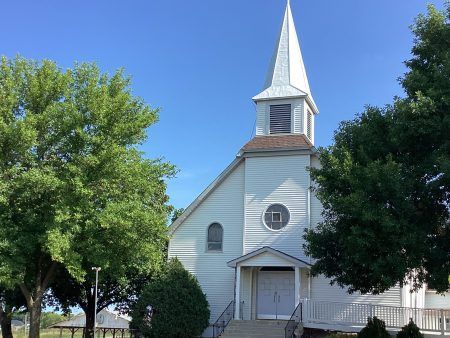 A white church with a tall steeple, a round window, and a front entrance, framed by two large green trees under a blue sky.