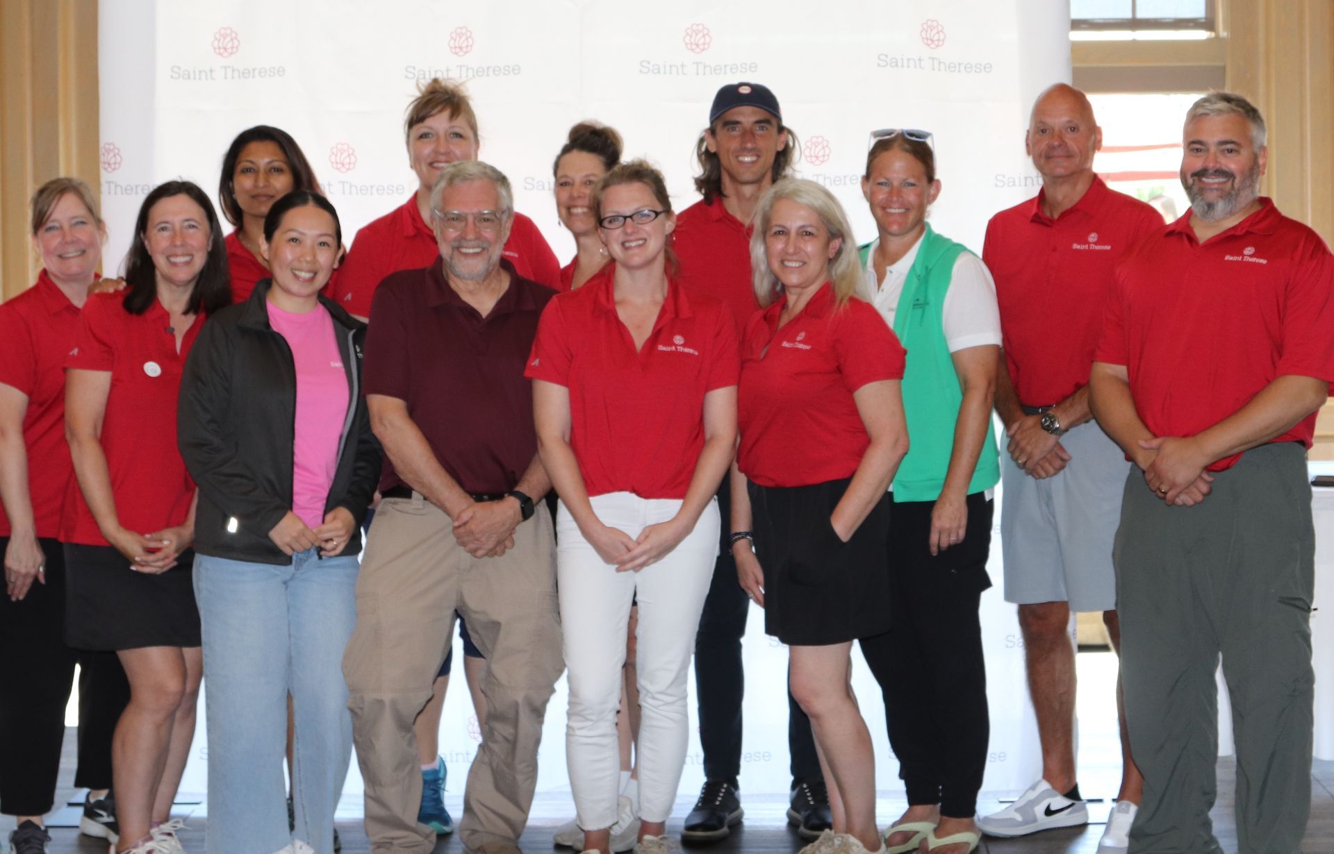 A group of fifteen people in red shirts and casual attire posing for a photo in front of a white branded backdrop.