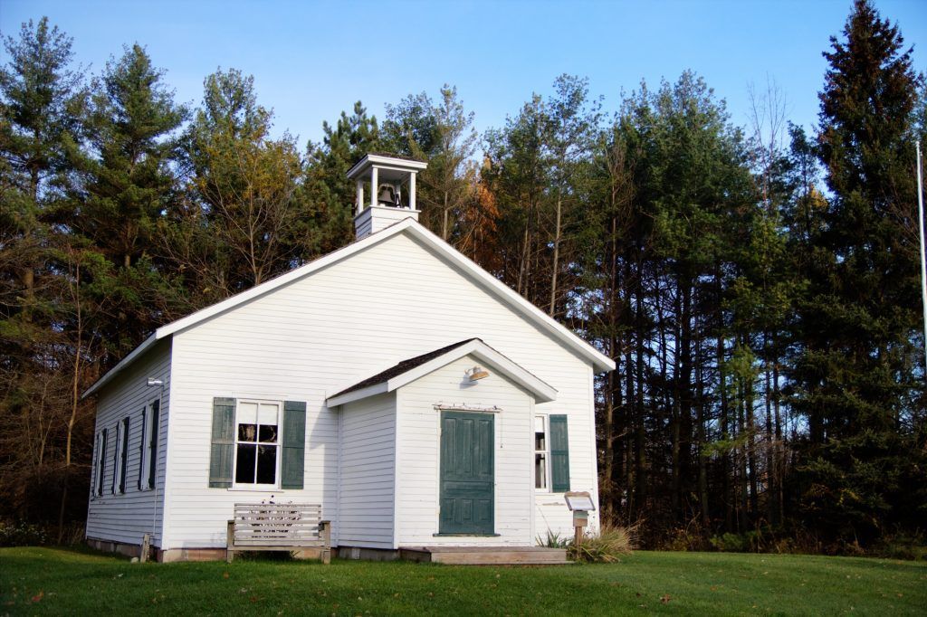 A small white, one-room schoolhouse with a bell tower stands in a clearing in front of a dense forest.