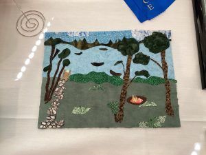A textile art scene showing a campsite with a fire, trees, rocky ground, and mountains under a blue sky.